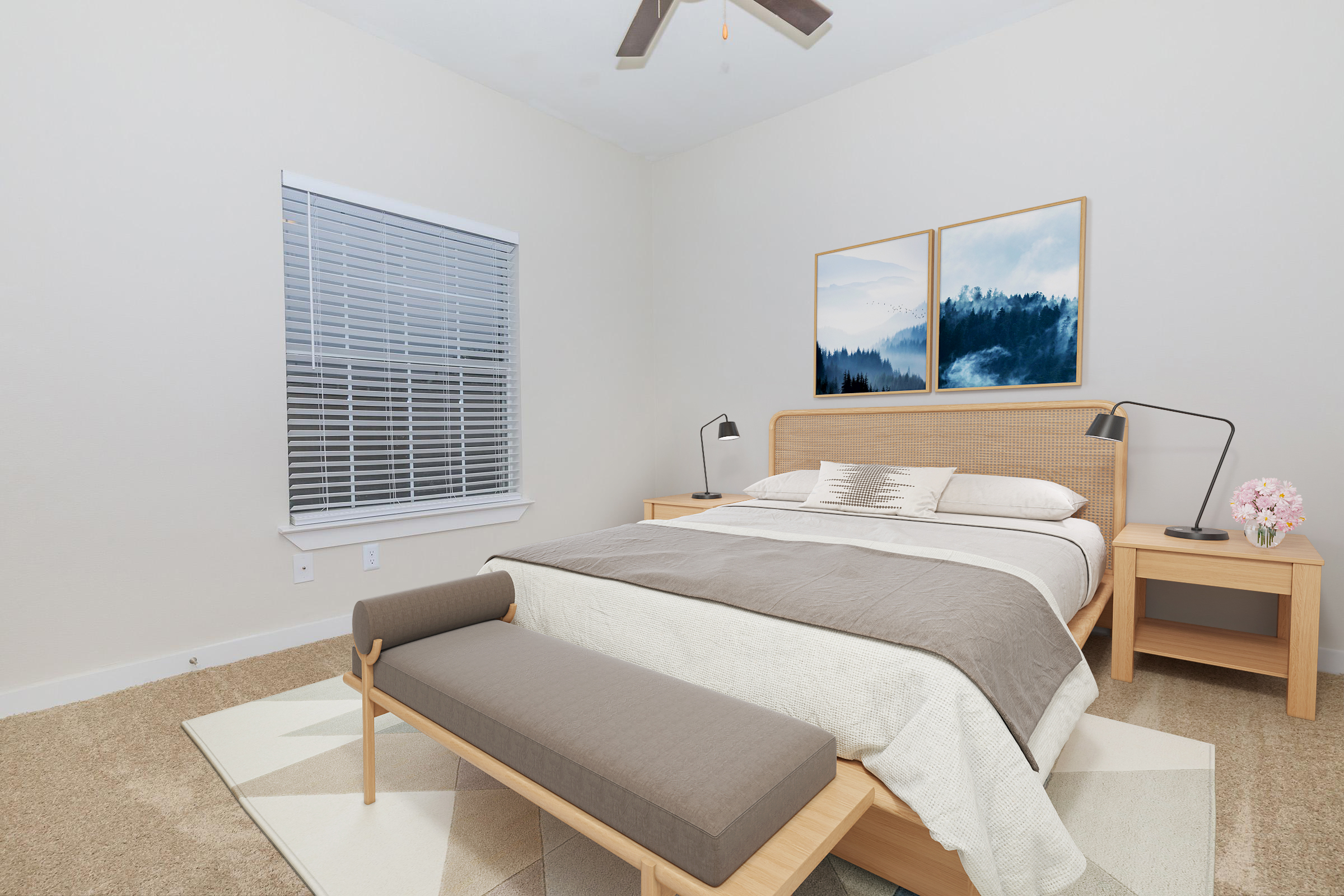 A well-lit, modern bedroom featuring a wooden bed frame with a light gray bedspread, two framed landscape paintings on the wall, a bench at the foot of the bed, and bedside tables with lamps. A window with blinds allows natural light, and a beige rug covers the floor.