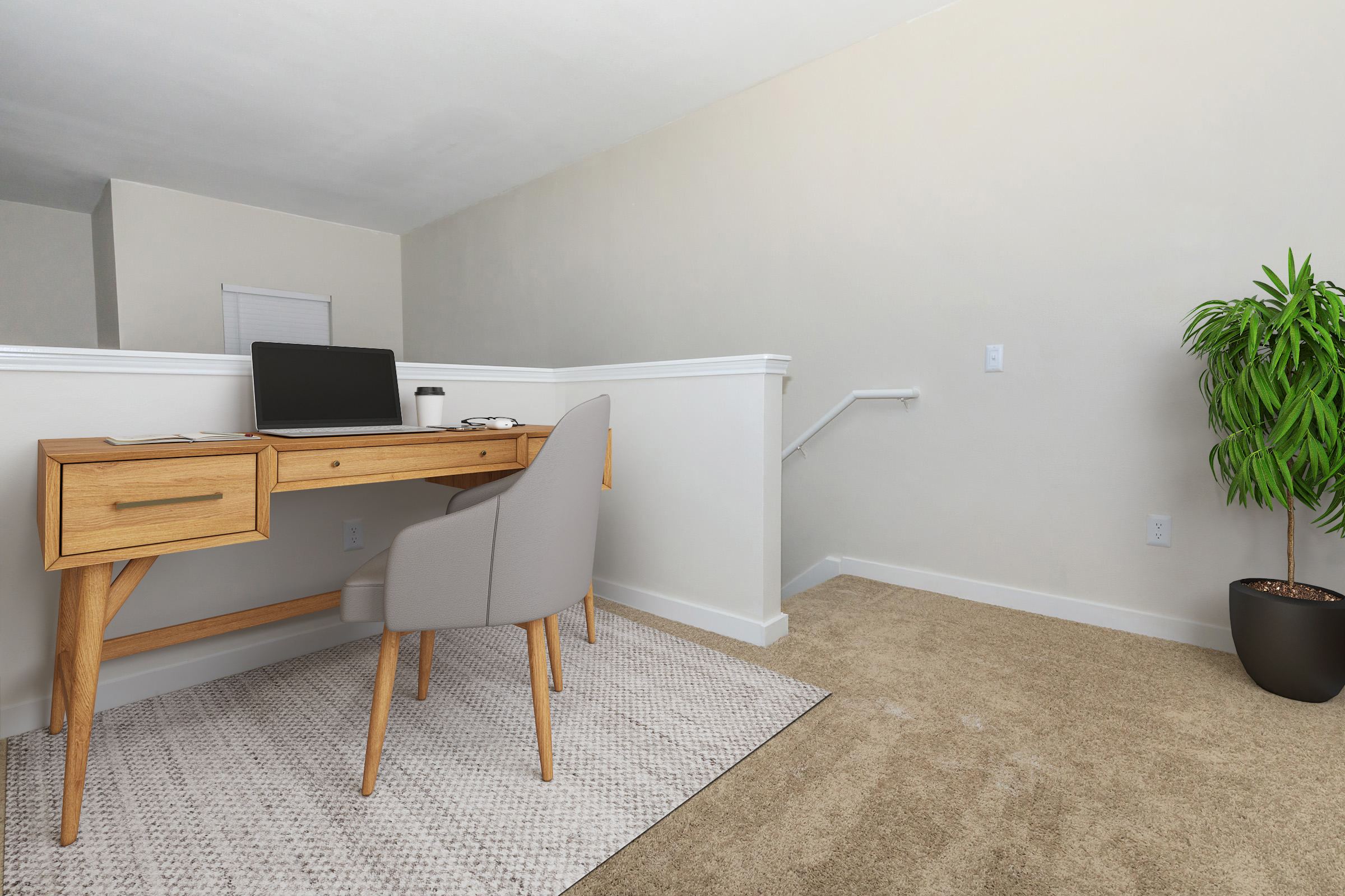 A minimalist home office setup featuring a wooden desk with a laptop, a coffee cup, and a gray chair. A light-colored wall and carpet complement the space. A green potted plant adds a touch of nature, while a staircase leads to another area in the background.