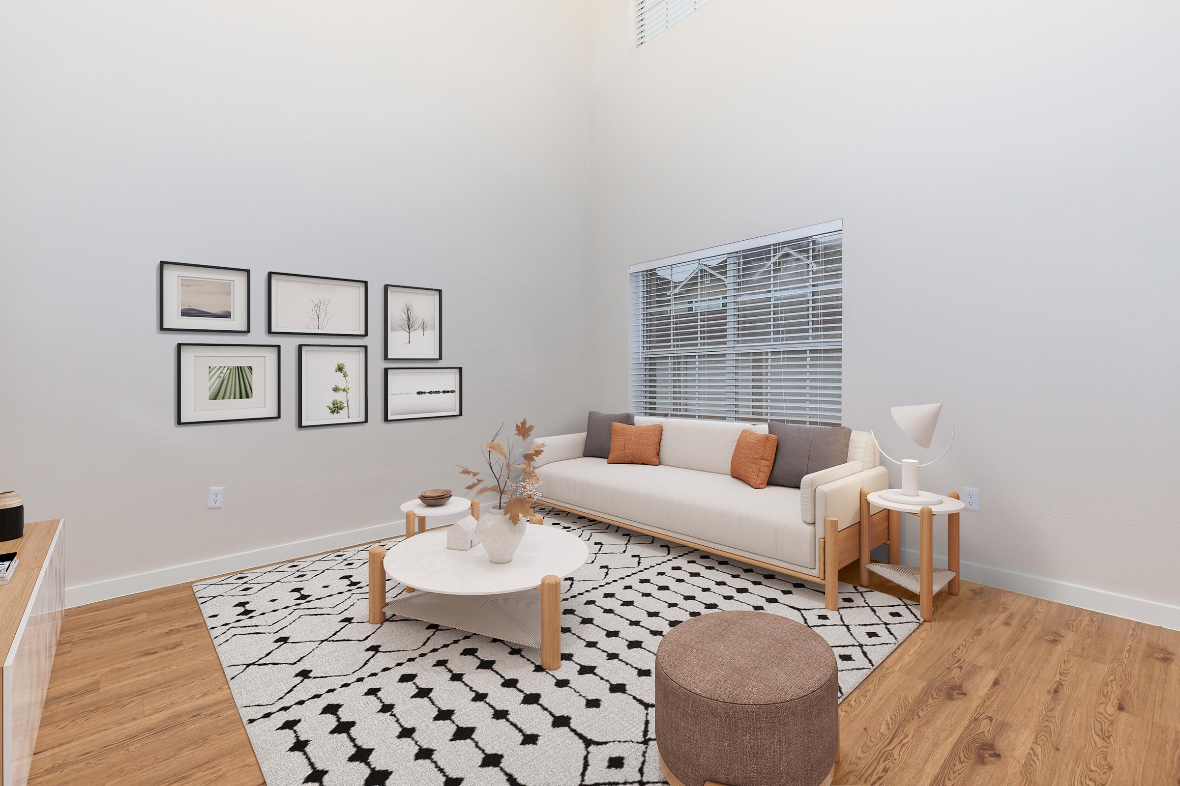 A modern living room featuring a white sofa with orange cushions, a round coffee table, and a cozy brown ottoman. The walls are decorated with framed artwork, while a large window allows natural light to fill the space. The floor is wooden, and a patterned area rug adds warmth to the room.