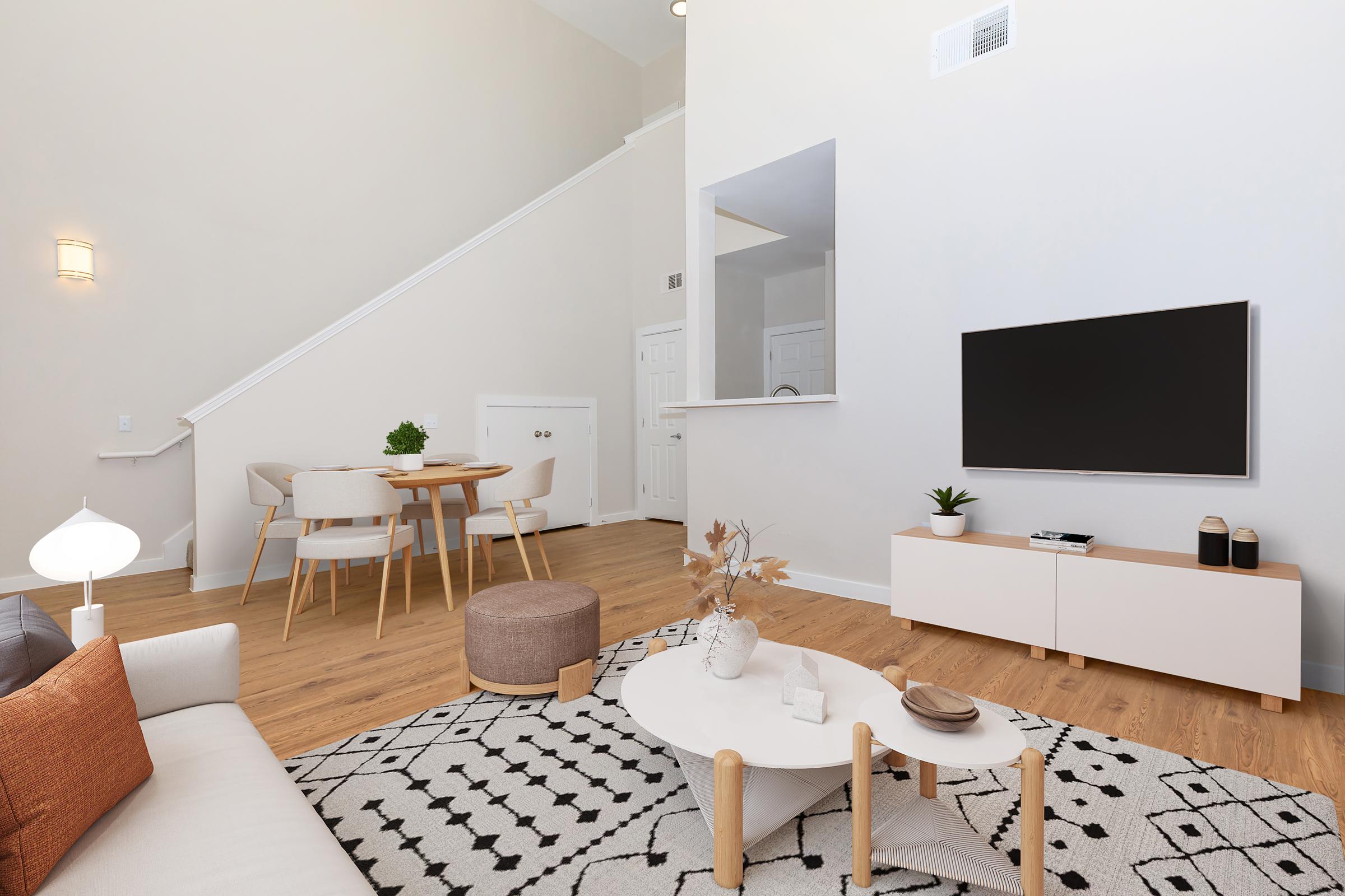 A modern living room featuring a light color palette. There’s a comfortable sofa with orange and white cushions, a round ottoman, and a stylish coffee table with decorative items. A flat-screen TV is mounted on the wall opposite a dining area with a wooden table and four chairs. Light wooden flooring and warm lighting create a cozy atmosphere.