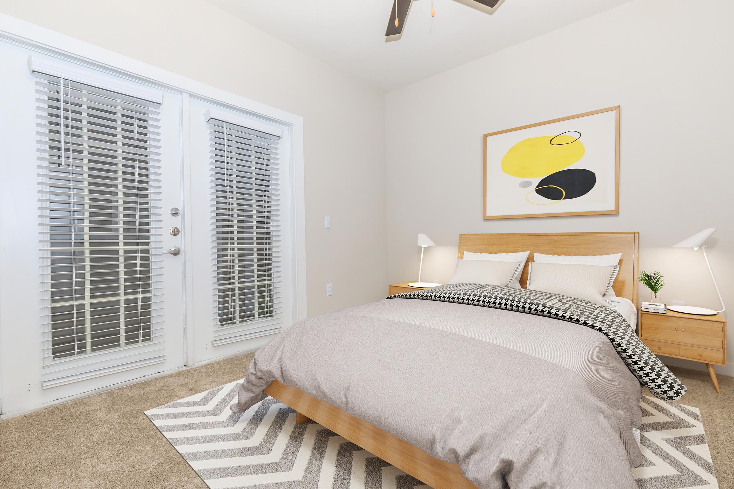 A cozy bedroom featuring a queen-sized bed with a light gray blanket, two bedside tables with lamps, and a small plant. There are large windows with white blinds and a modern piece of wall art above the bed. The floor is carpeted with a striped area rug.