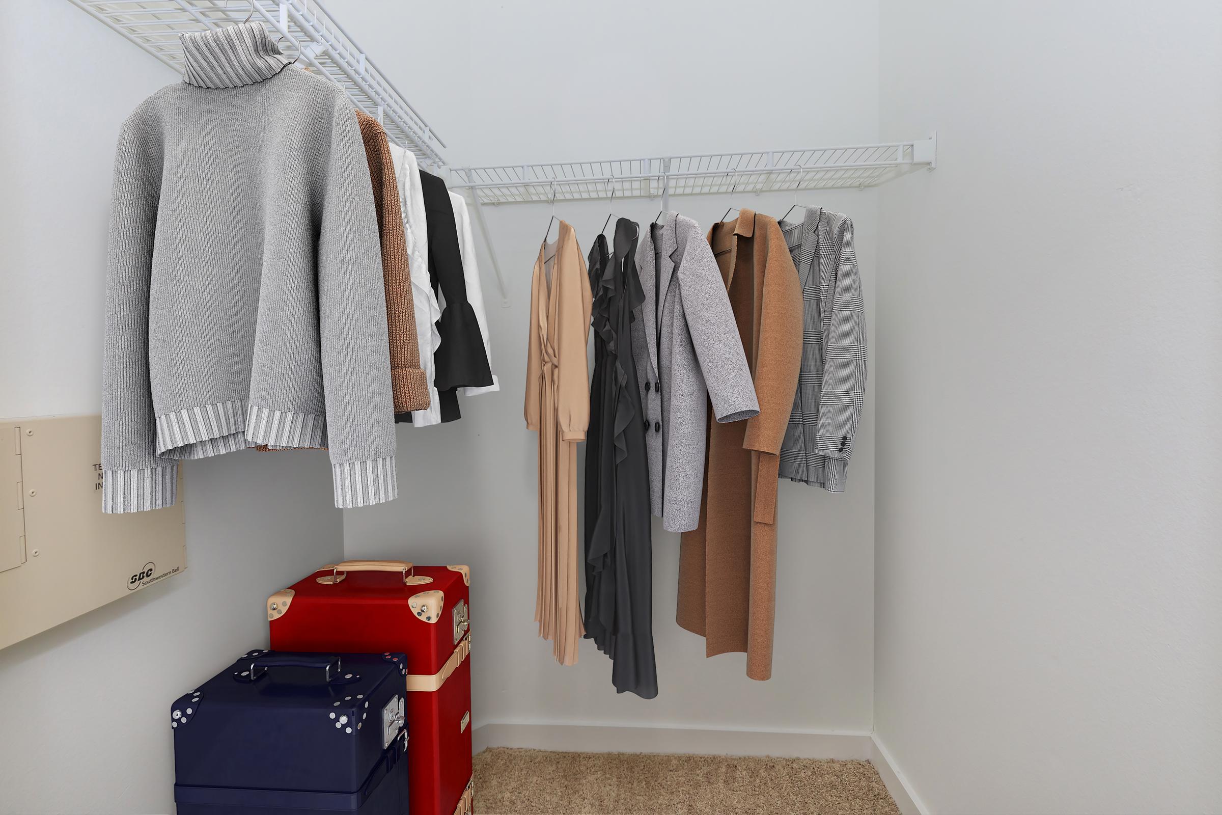 A neatly organized closet featuring a variety of clothing, including sweaters, dresses, and suits hanging on a wire rack. Two vintage suitcases in red and blue are positioned on the floor, complementing the clean and tidy appearance of the space.