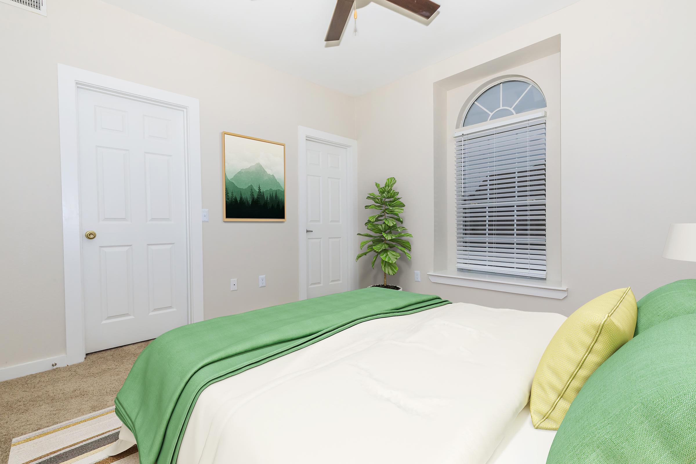 A cozy bedroom featuring a green bedspread with yellow pillows, a potted plant in the corner, and a large window with closed blinds. The walls are a soft beige, and there's a piece of mountain-themed art on the wall. A ceiling fan adds to the warm ambiance, enhancing the inviting atmosphere of the space.