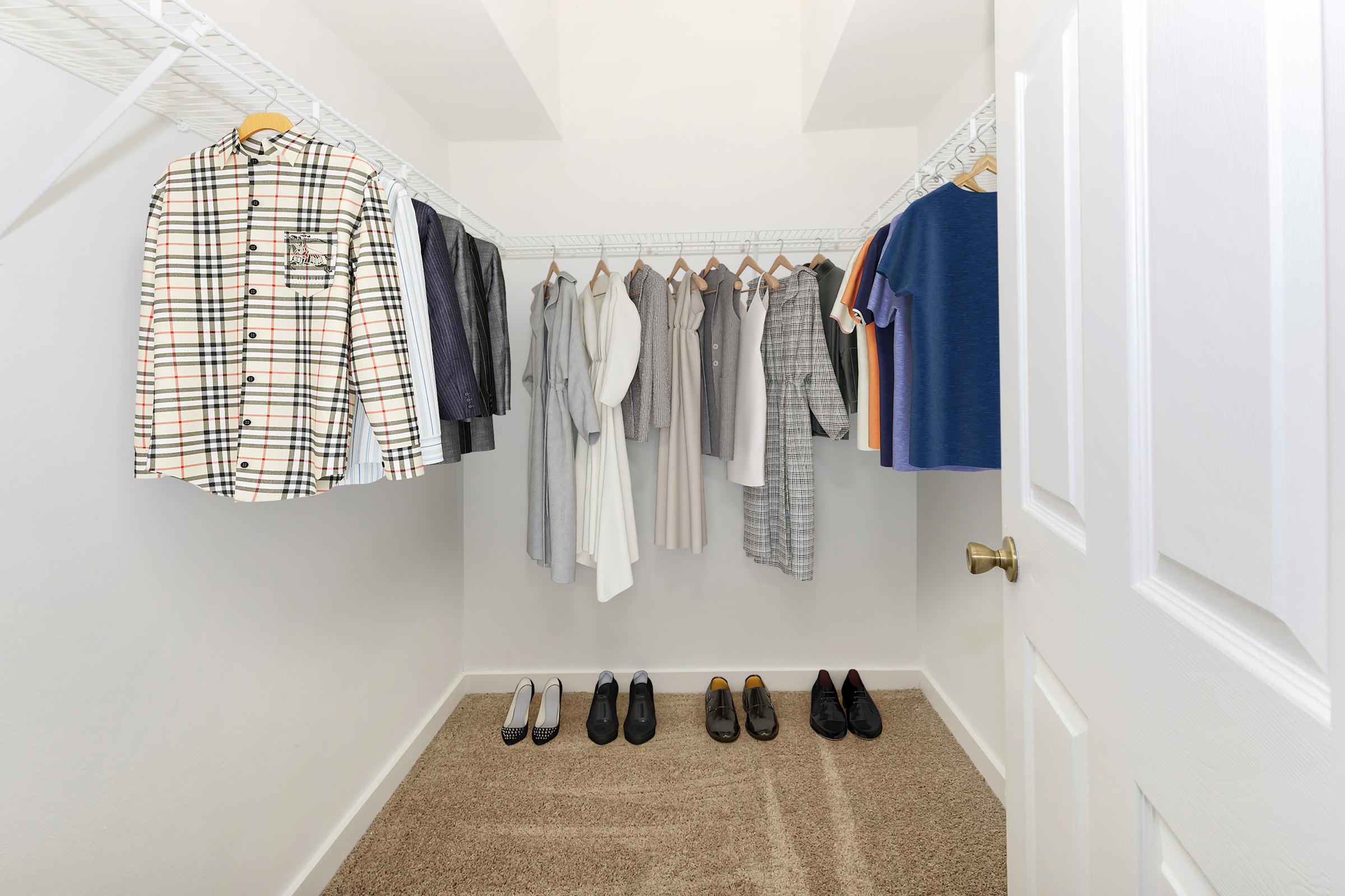 A neatly organized closet showcasing a variety of clothing items. On the left, a plaid shirt hangs alongside several other garments in neutral tones, including dresses and blouses. Below, there are pairs of shoes, including heels and dress shoes, arranged on a carpeted floor. The closet has white wire shelving and a closed door.