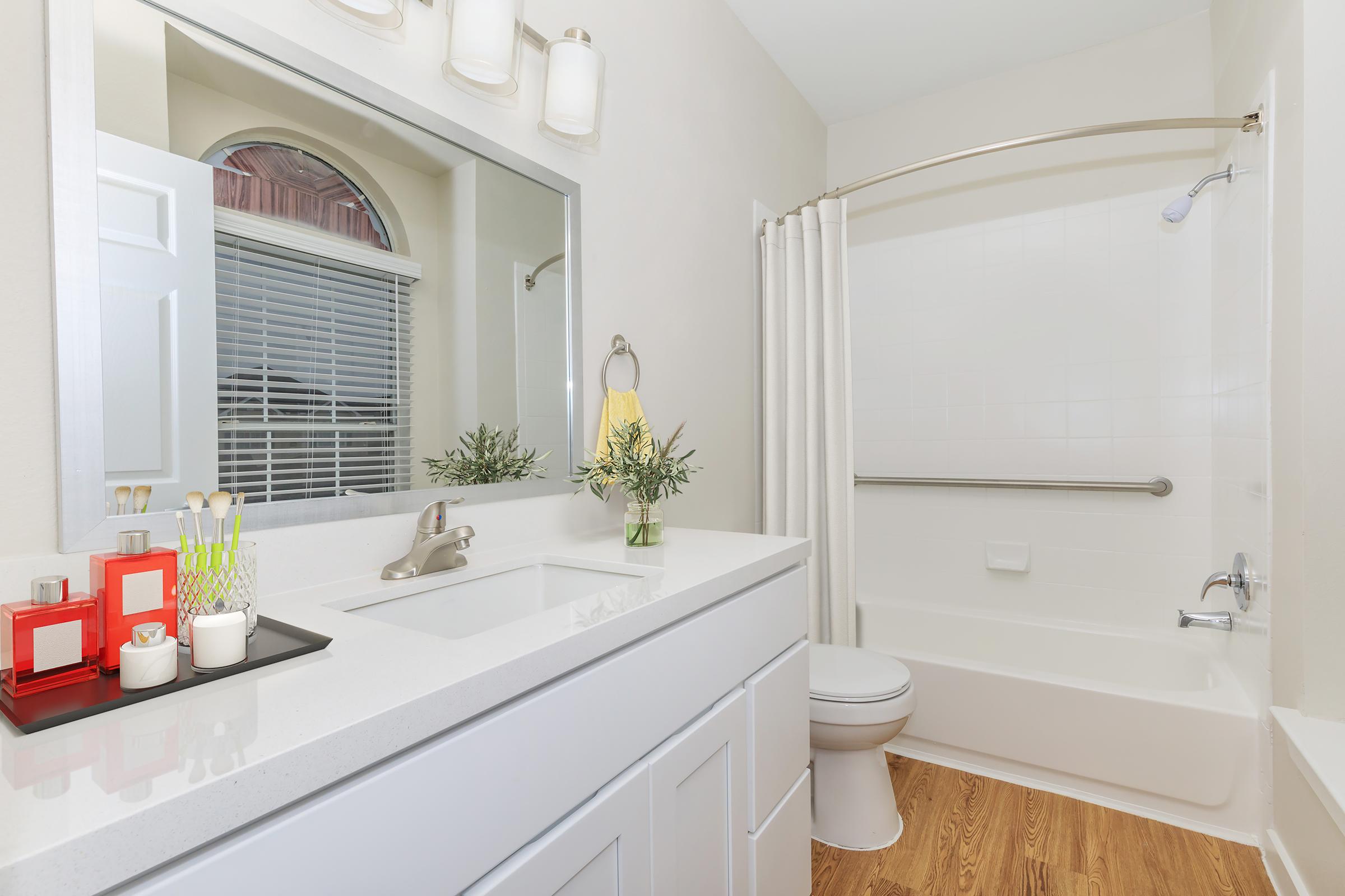 A modern bathroom featuring a white vanity with a sink, a large mirror above, and organized toiletries. There’s a bathtub with a shower curtain, a toilet, and a window allowing natural light. The wooden floor adds warmth to the space, complemented by decorative elements like plants and candles on the counter.