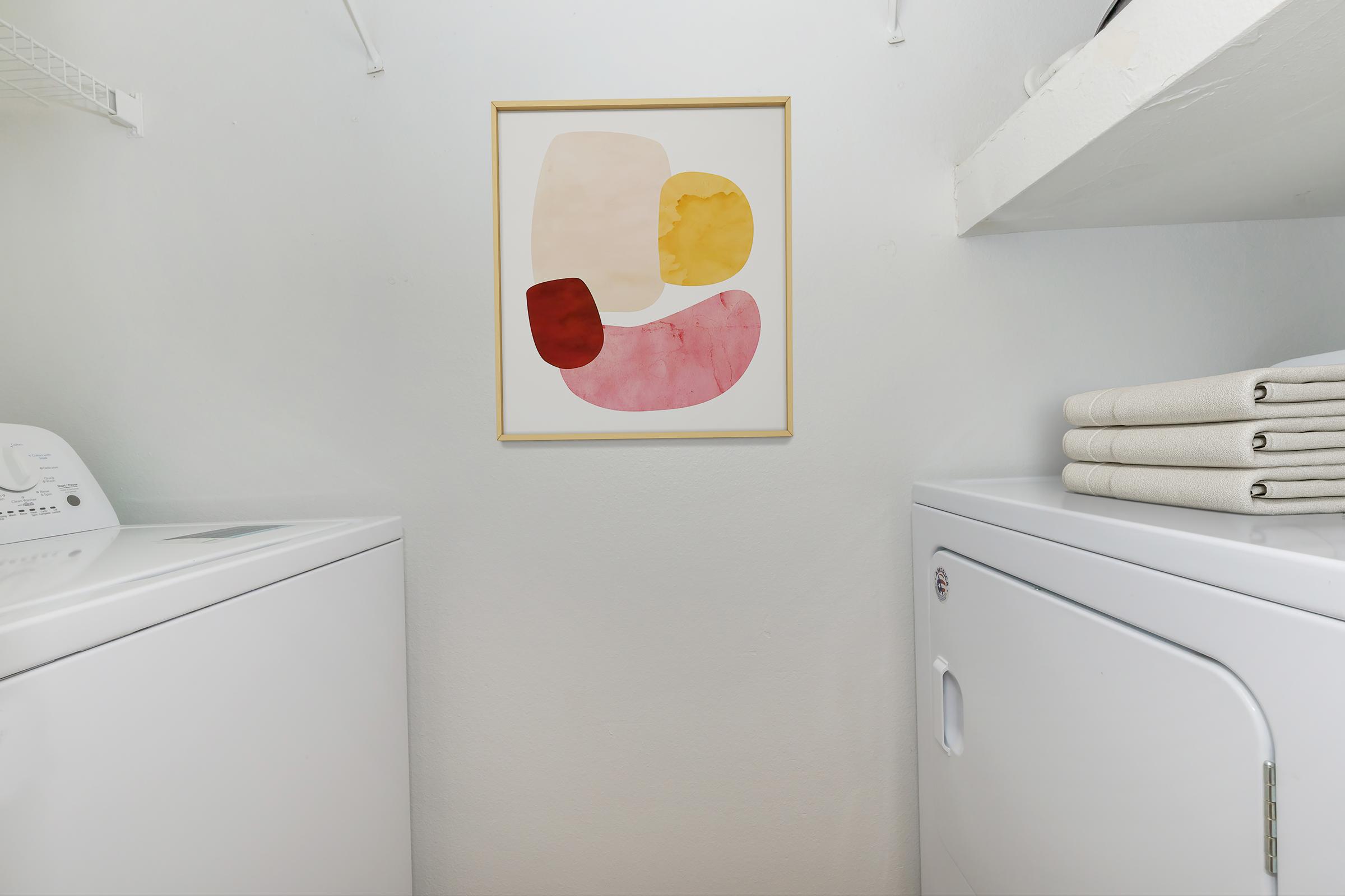 A bright and modern laundry room featuring a white washing machine and dryer. On the wall, there is a framed abstract art piece with shapes in soft pink, red, and yellow hues. Neatly folded beige towels are stacked on a shelf above the appliances, contributing to the clean and organized atmosphere.