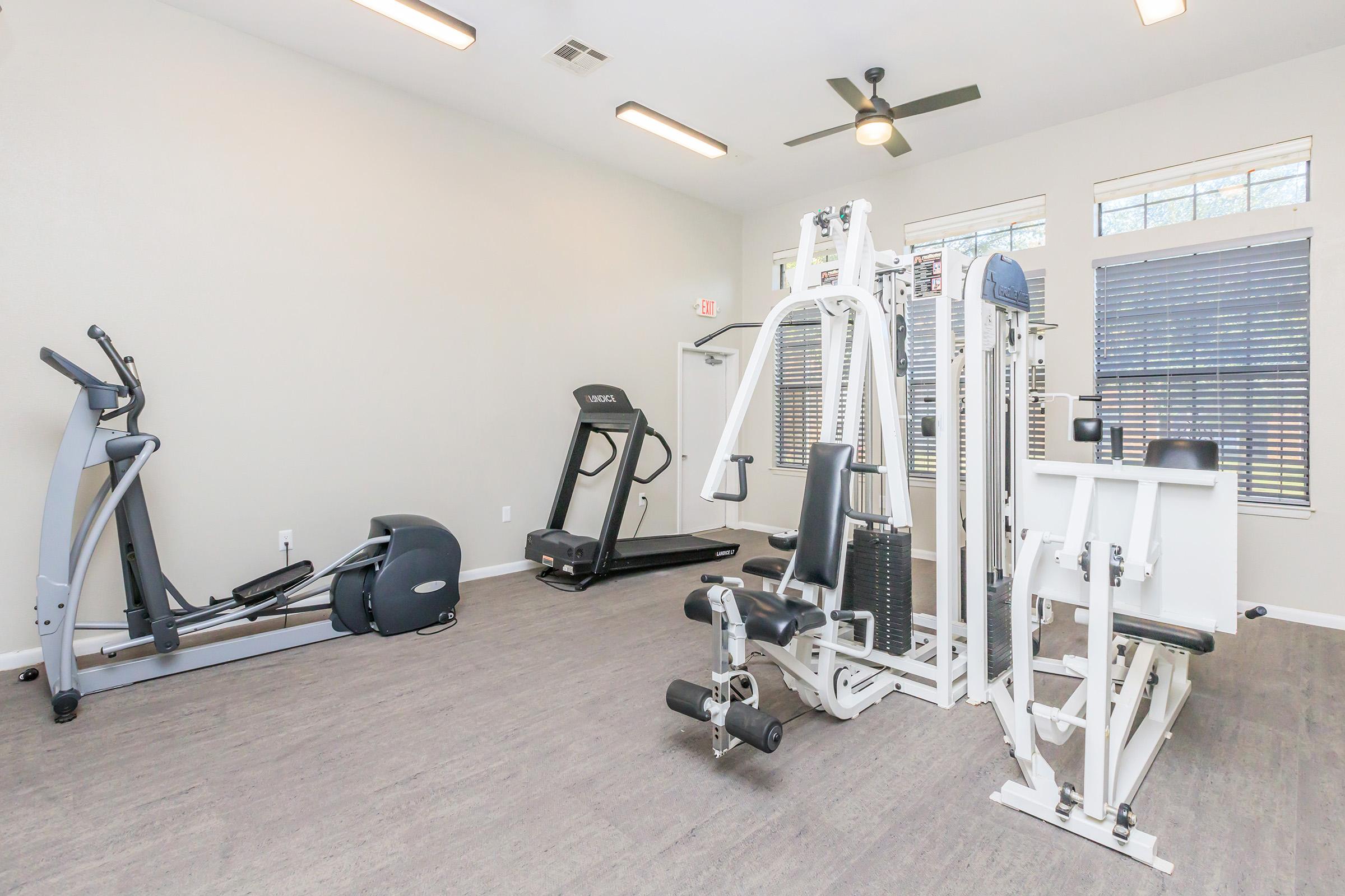 A well-lit gym area featuring various exercise equipment including a treadmill, elliptical machine, and a multi-station weight machine. The room has large windows for natural light and a neutral color scheme, creating an inviting space for workouts.