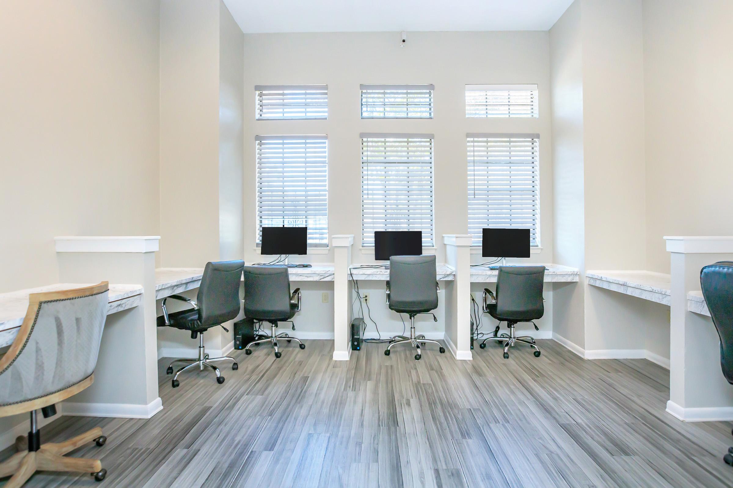 A modern workspace featuring four computer desks with chairs, positioned against a wall with large windows. The room has light-colored walls and wooden flooring, creating a bright and inviting atmosphere for work or study.