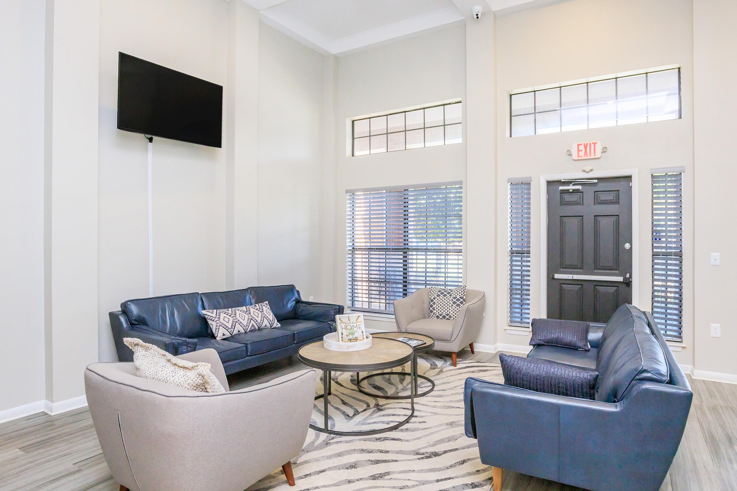 A modern lounge area featuring two blue sofas and a light-colored armchair arranged around a round coffee table. The room has light walls, large windows with blinds, and a flat-screen TV mounted on the wall. A decorative area rug adds texture to the floor.