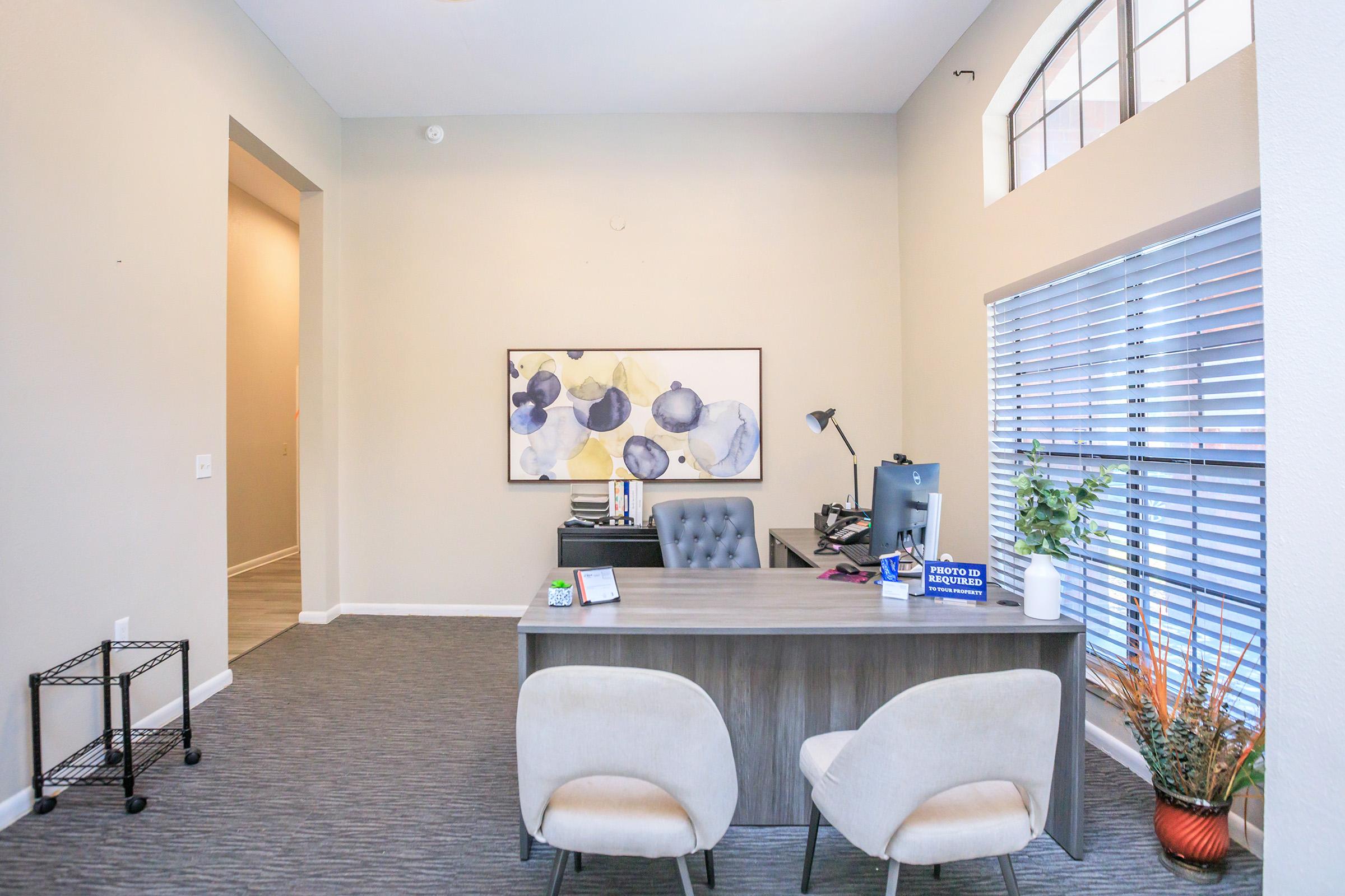 A modern office space featuring a wooden desk with a computer, a comfortable chair, and a decorative plant. The walls are painted light beige, with a large window featuring blinds. A piece of abstract art hangs above the desk, and there’s a black cart in the corner. The room is well-lit and inviting.