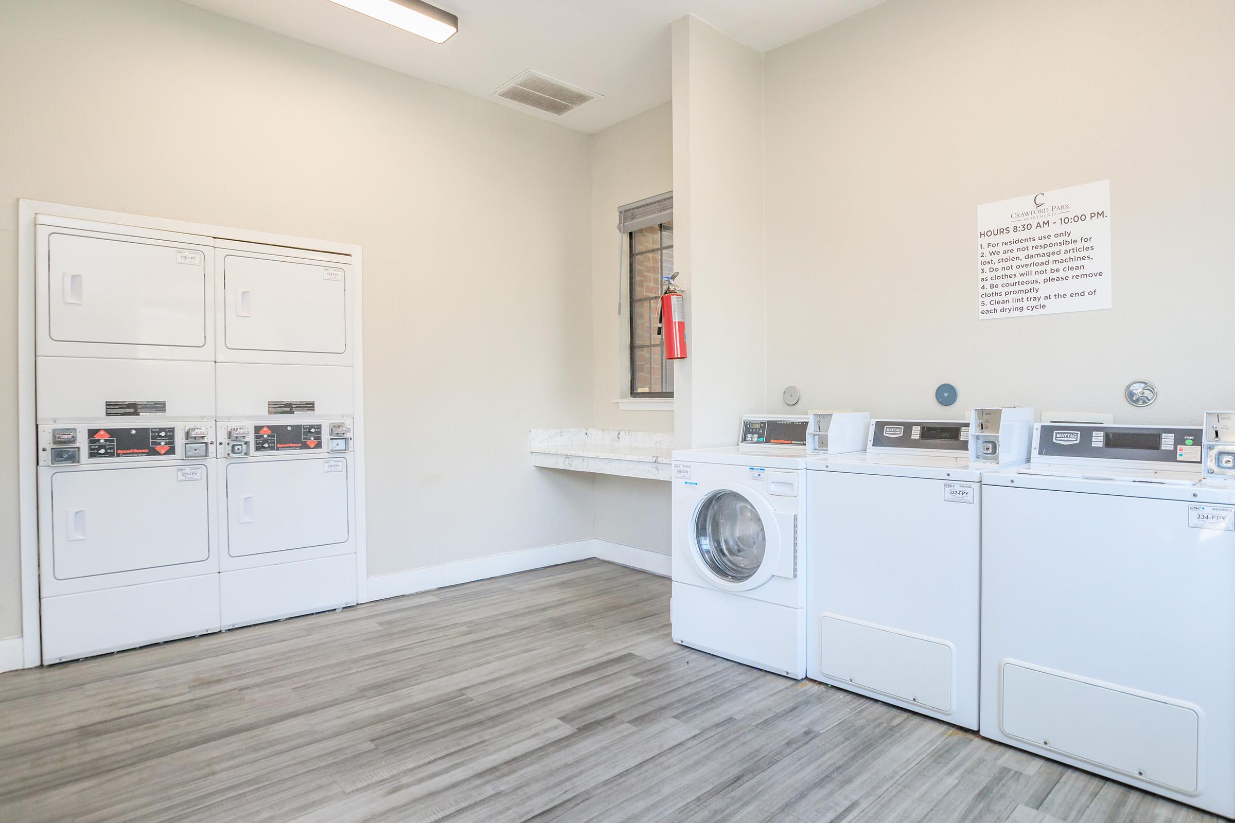 A clean, modern laundry room featuring white stackable washing machines and dryers. A countertop is available for folding clothes. The walls are painted a light color, and there's a notice on the wall with laundry room rules. The floor has a wood-like finish, creating a bright and inviting space.