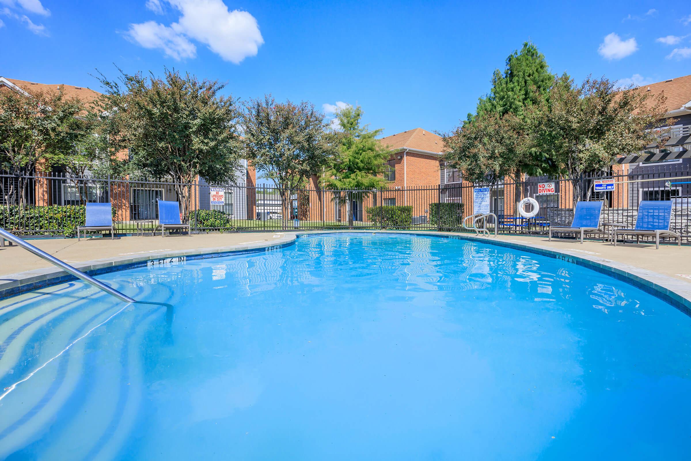 A tranquil swimming pool surrounded by lush greenery and lounge chairs. The clear blue water reflects a bright sky with scattered clouds, and a fenced area adds privacy to the pool space. Trees line the perimeter, creating a relaxing atmosphere for visitors.