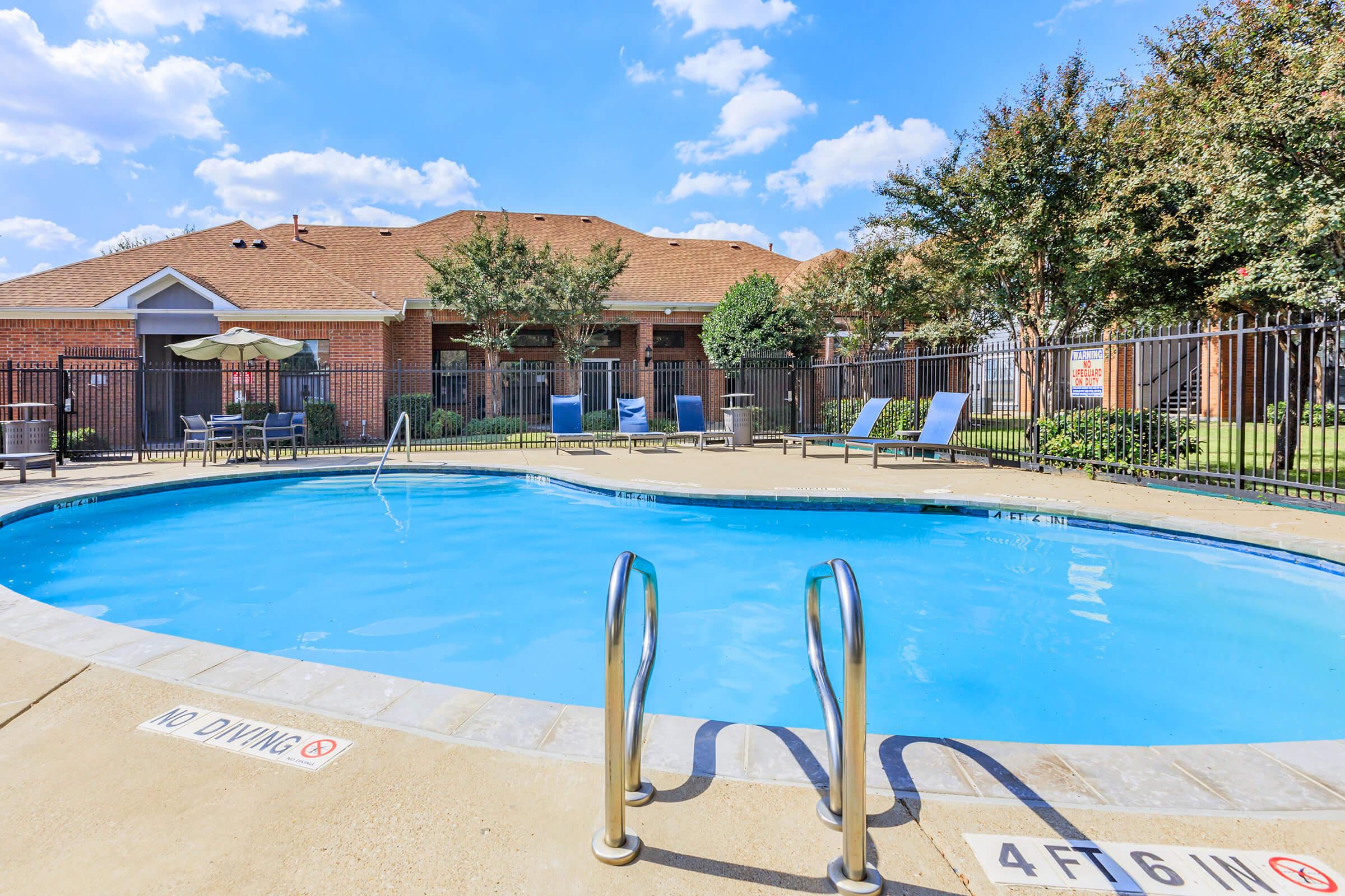 A sparkling blue swimming pool with a shallow end, surrounded by a fenced area. Lounge chairs are positioned nearby under shaded umbrellas. In the background, a residential building is visible, with trees and a clear blue sky dotted with fluffy clouds above.