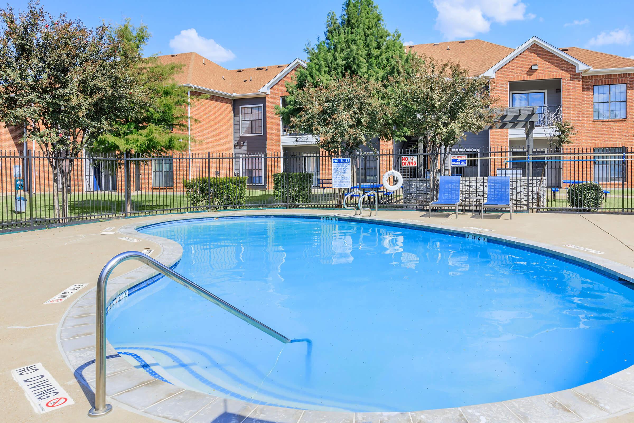 A serene outdoor swimming pool with a clear blue surface, surrounded by a fenced area and neatly landscaped greenery. In the background, there are brick apartment buildings, and a few patio chairs are lined up beside the pool. The scene is bright and inviting under a clear blue sky.