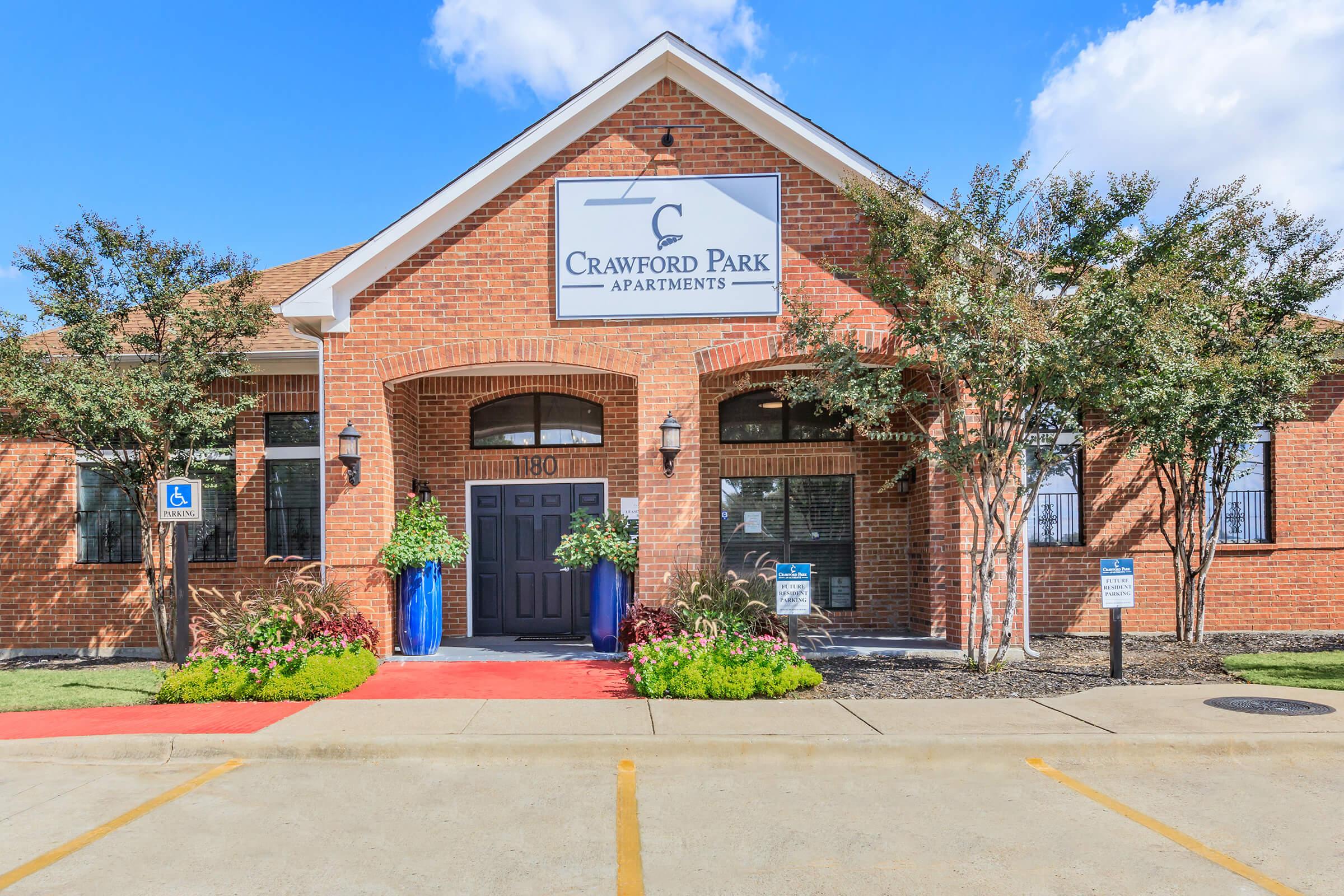 A brick apartment building with the sign "Crawford Park Apartments" above the entrance. The entrance features glass double doors, large windows, and decorative plants on either side. A red pathway leads up to the door, and there are signs indicating accessible parking. The sky is partly cloudy.