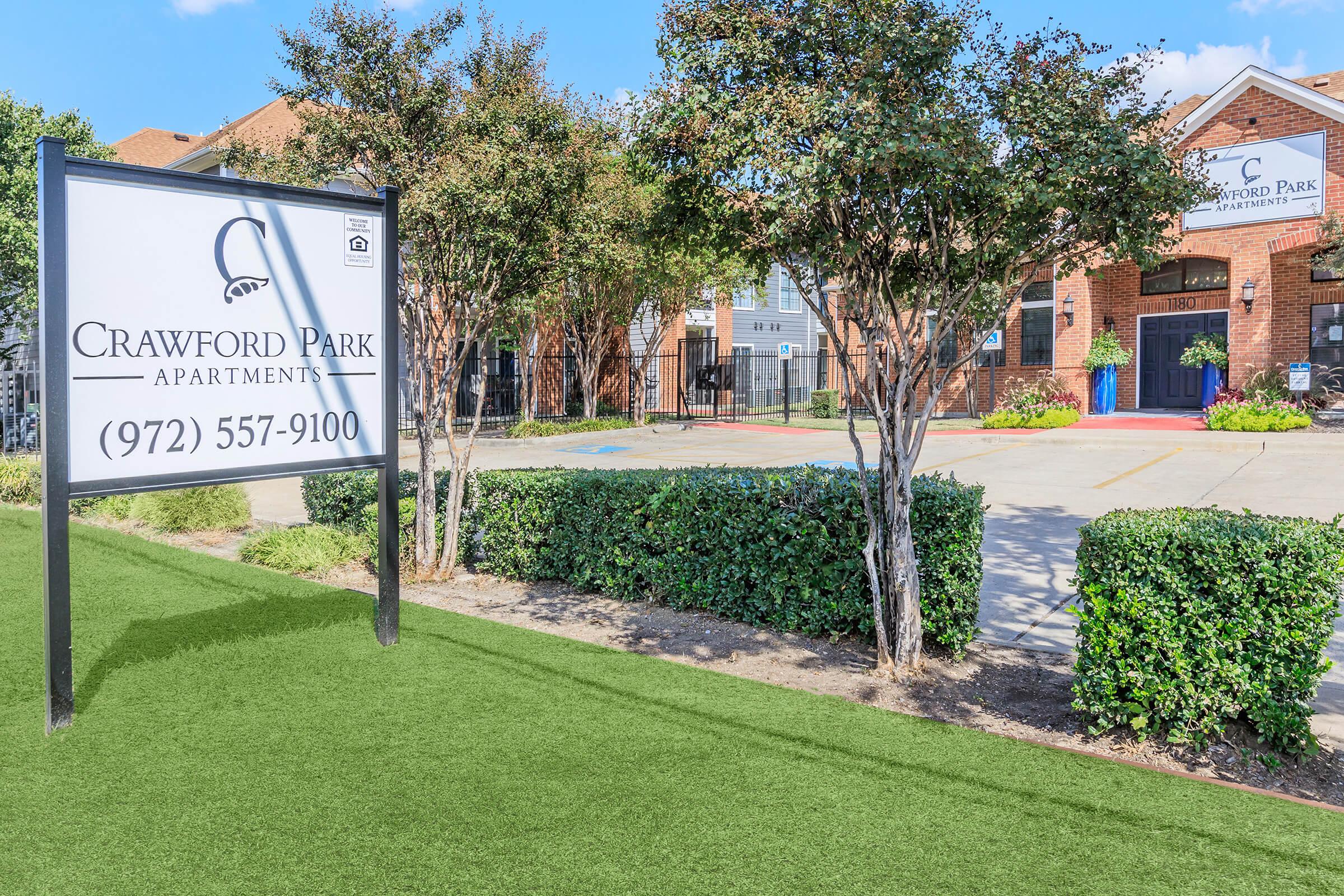 Sign for Crawford Park Apartments with the phone number (972) 557-9100 in front of a landscaped area. The apartment complex features a brick exterior with trees and green grass, under a clear blue sky.