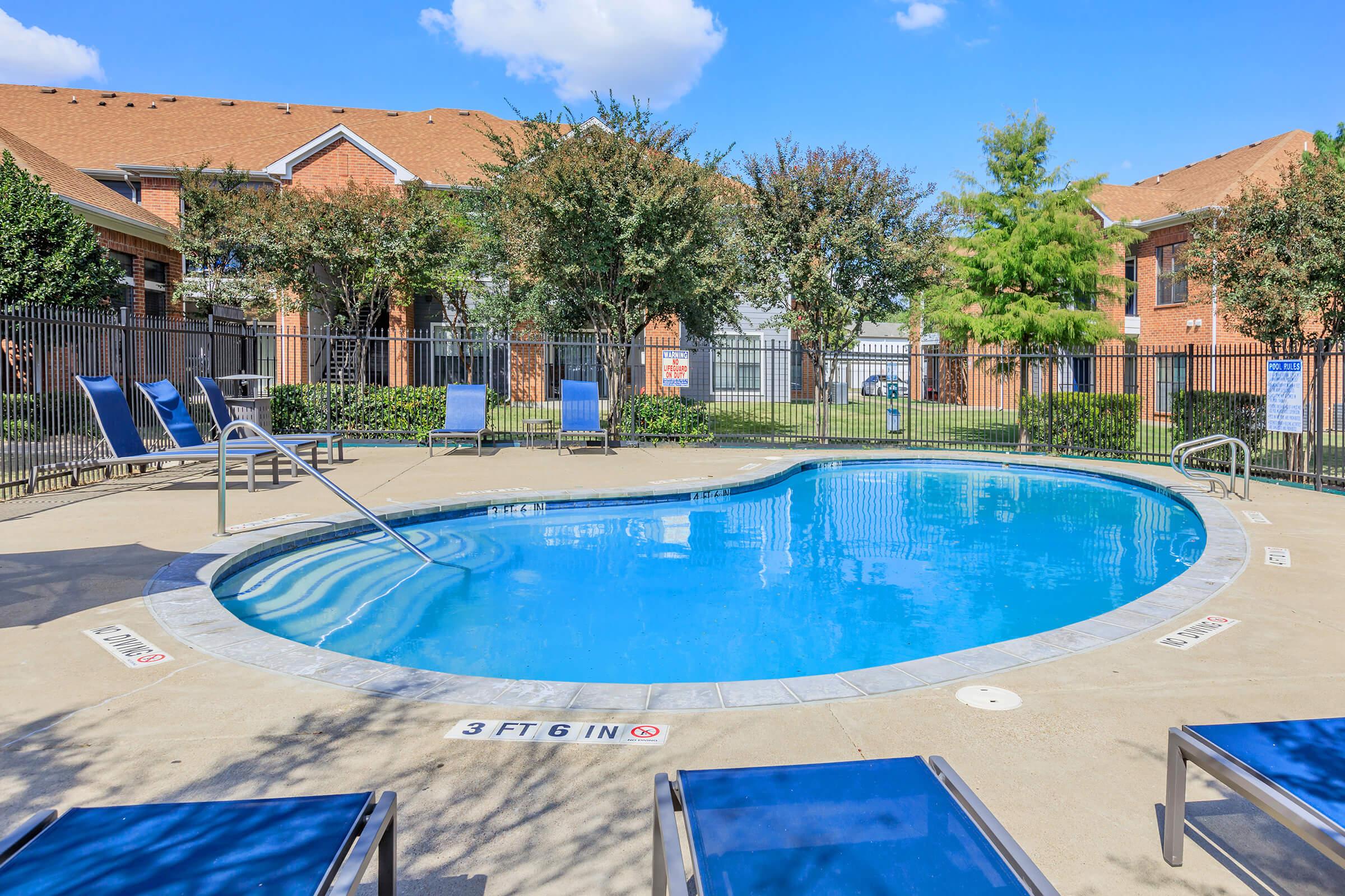 A clear blue swimming pool surrounded by lounge chairs, with a shallow area marked at 3 feet 6 inches. The pool area is enclosed by a black fence, and green trees and buildings can be seen in the background under a blue sky with fluffy white clouds.
