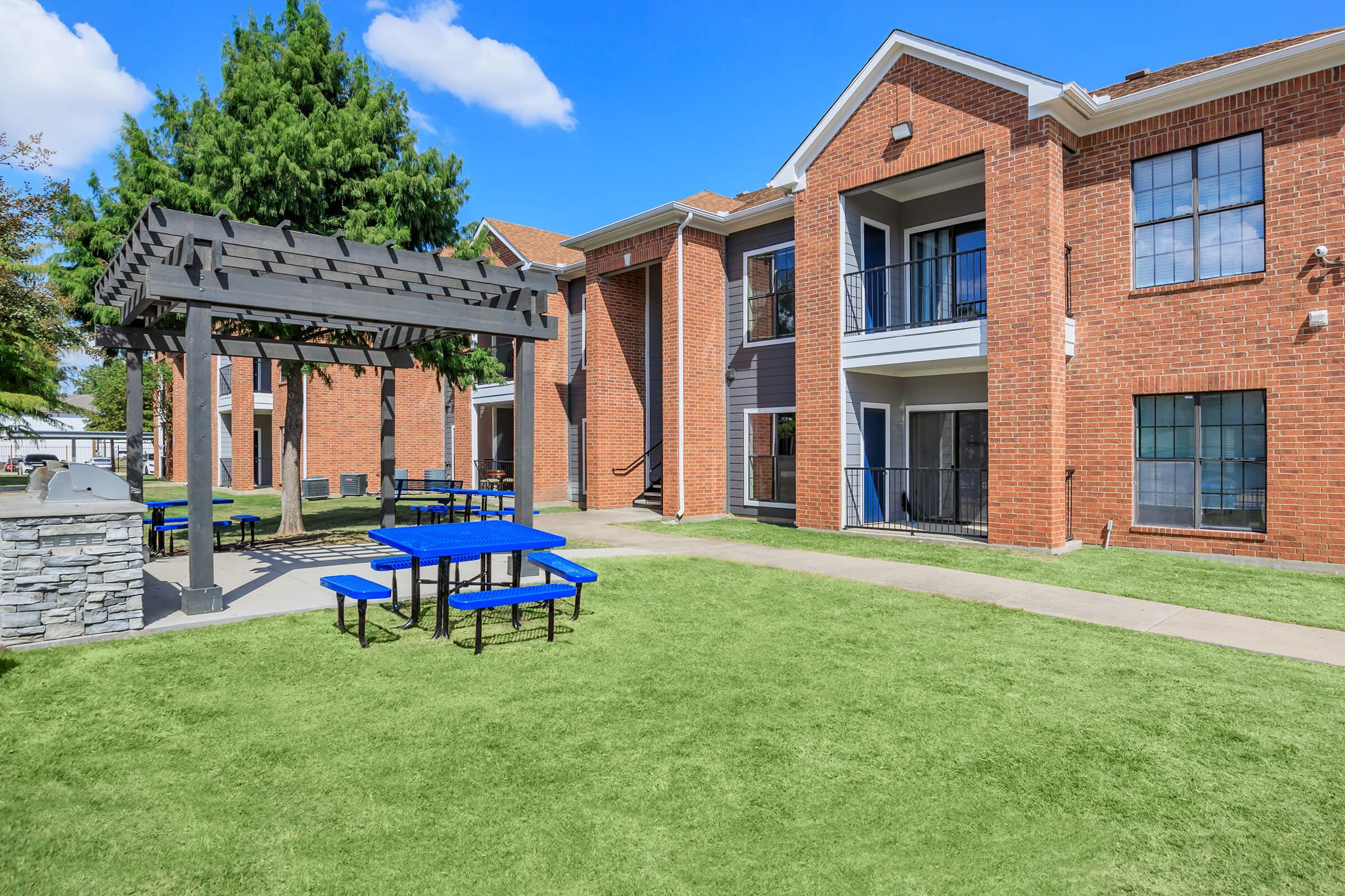 A well-maintained outdoor area featuring a grassy lawn, a pergola with a picnic table, and brick apartment buildings. The scene is set under a clear blue sky, creating a welcoming environment for residents and visitors.