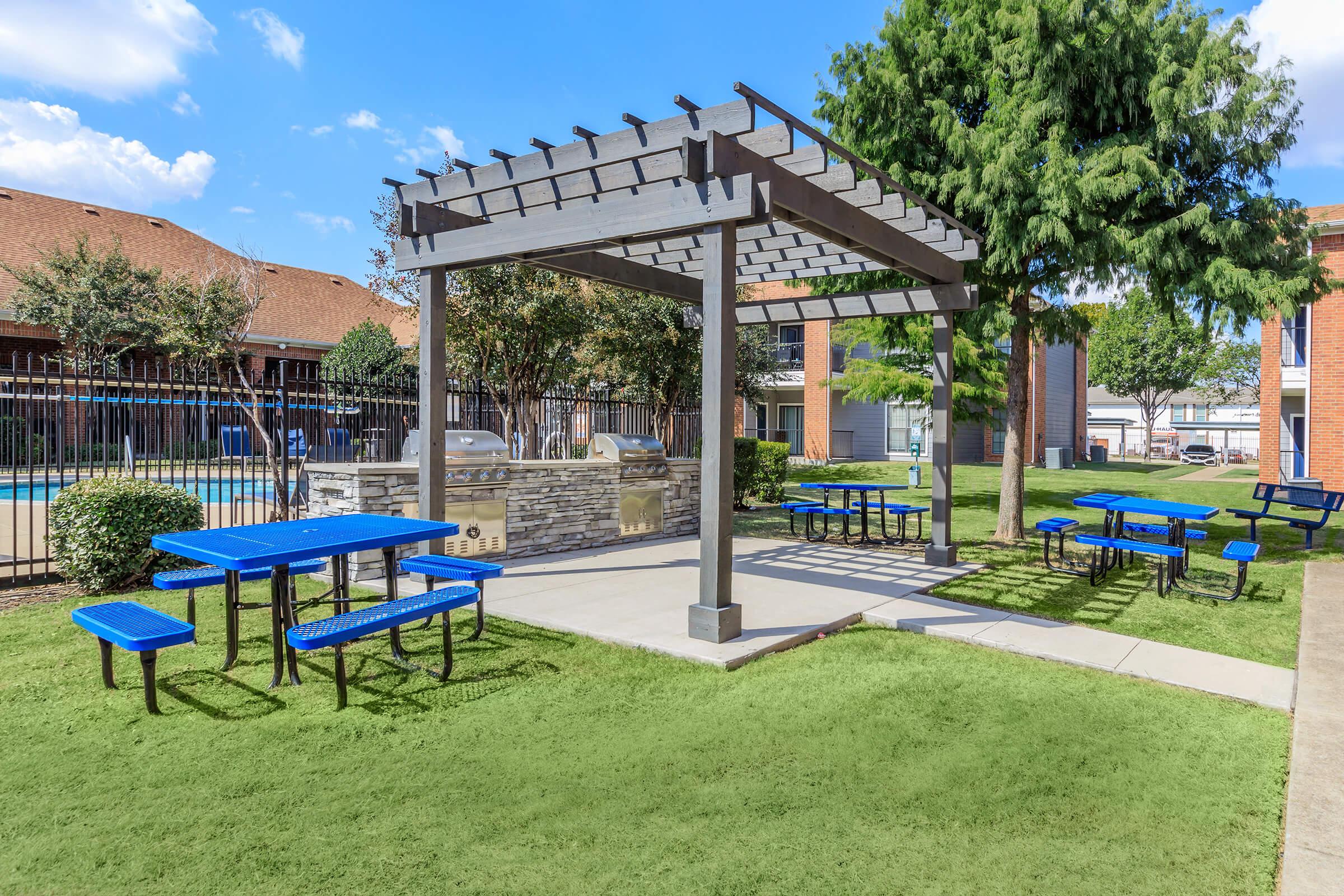 Outdoor gathering area featuring a pergola with benches and tables, surrounded by lush green grass and trees. In the background, a fenced pool area and brick buildings can be seen under a clear blue sky with a few clouds.