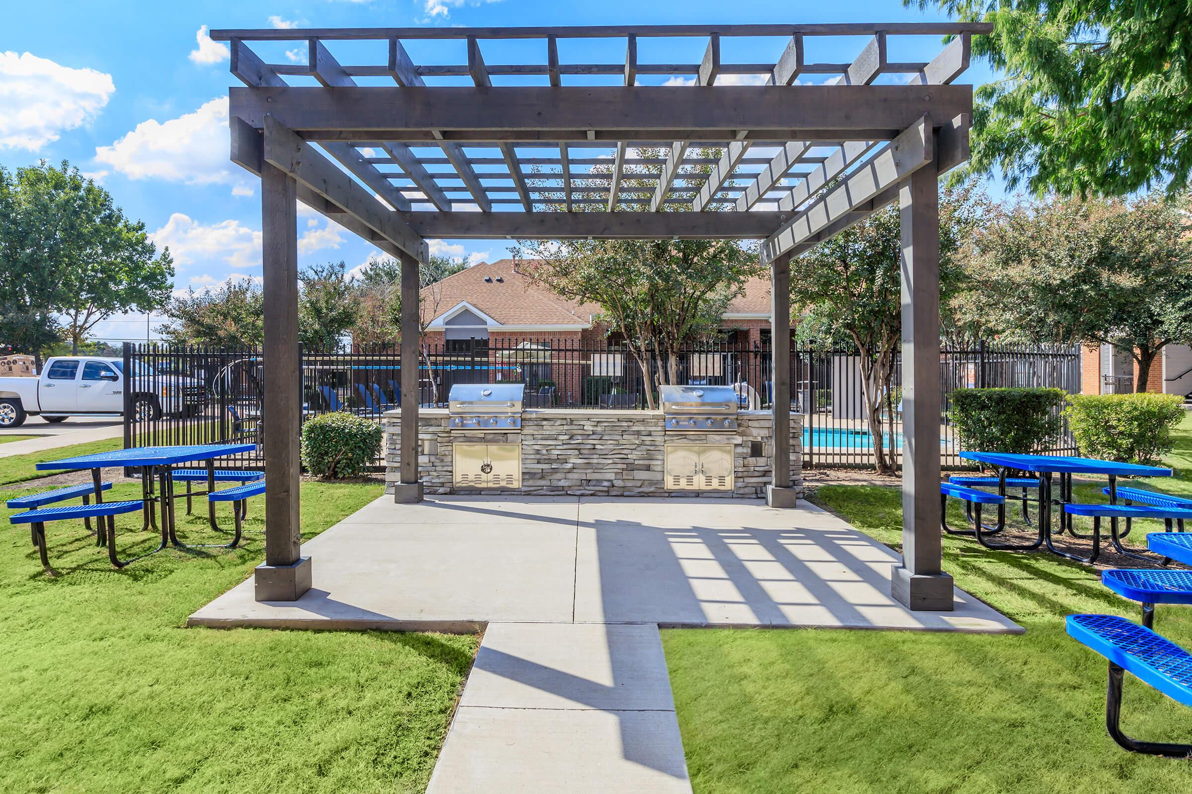 An outdoor grilling area featuring a wooden pergola with two barbecue grills, surrounded by picnic tables with blue tops. Lush green grass and trees frame the space, creating a welcoming atmosphere. In the background, a fenced pool area and residential buildings are visible under a clear blue sky.