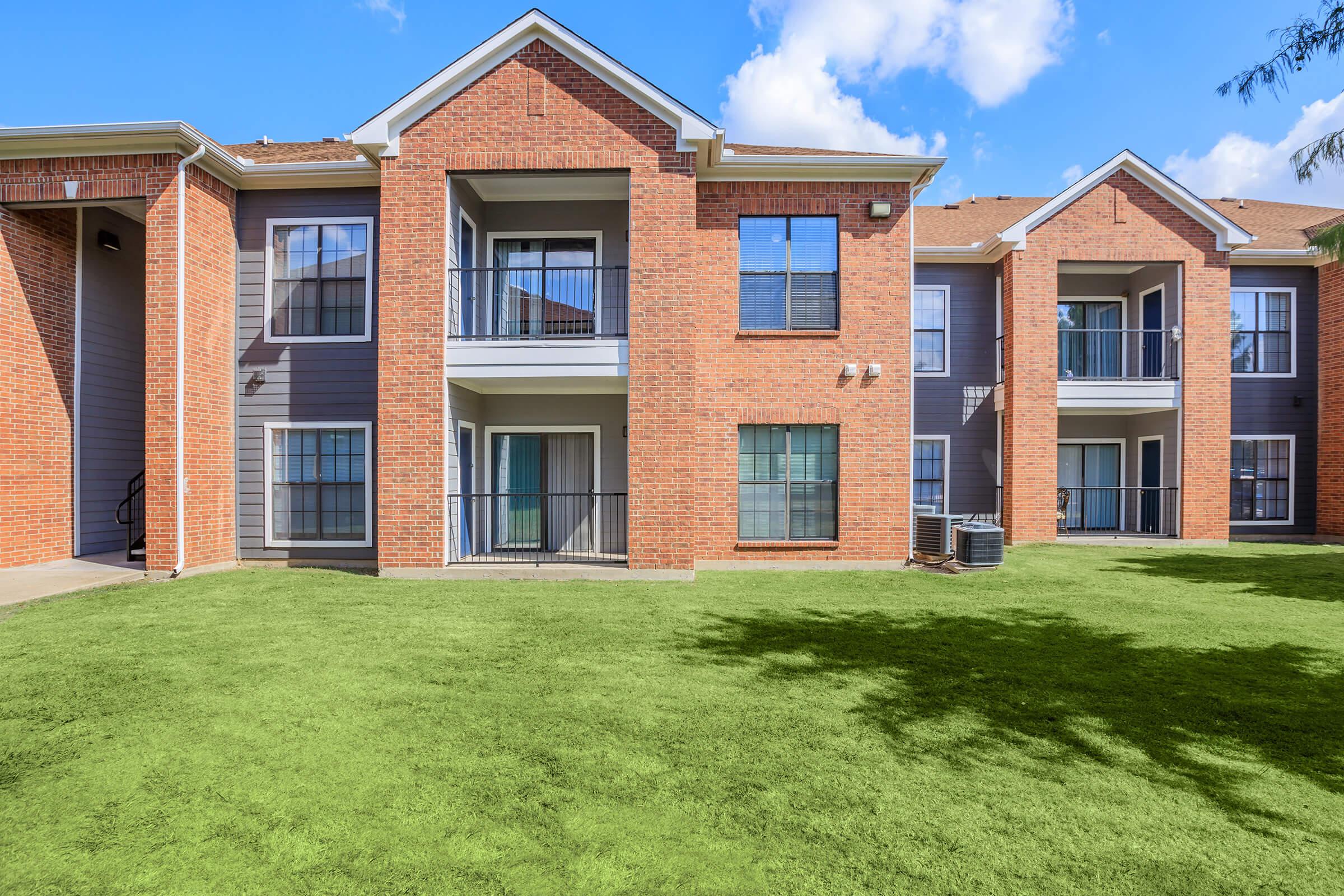 Two-story brick apartment buildings with balconies on the second floor, surrounded by a well-maintained green lawn. The sky is partly cloudy, adding a bright and inviting atmosphere to the setting.