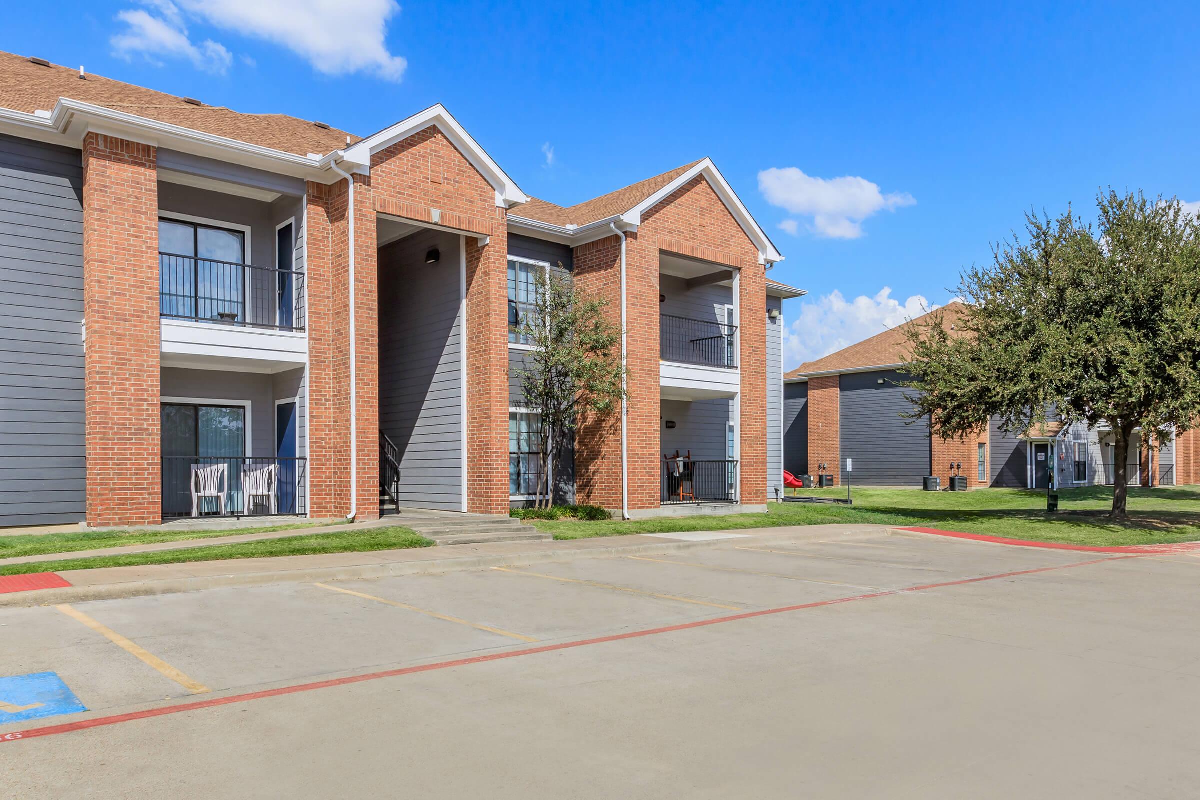 Two apartment buildings with brick and gray siding, featuring balconies. A clear blue sky with a few clouds overhead. The foreground shows a parking lot with marked spaces, and well-maintained grass and trees nearby.