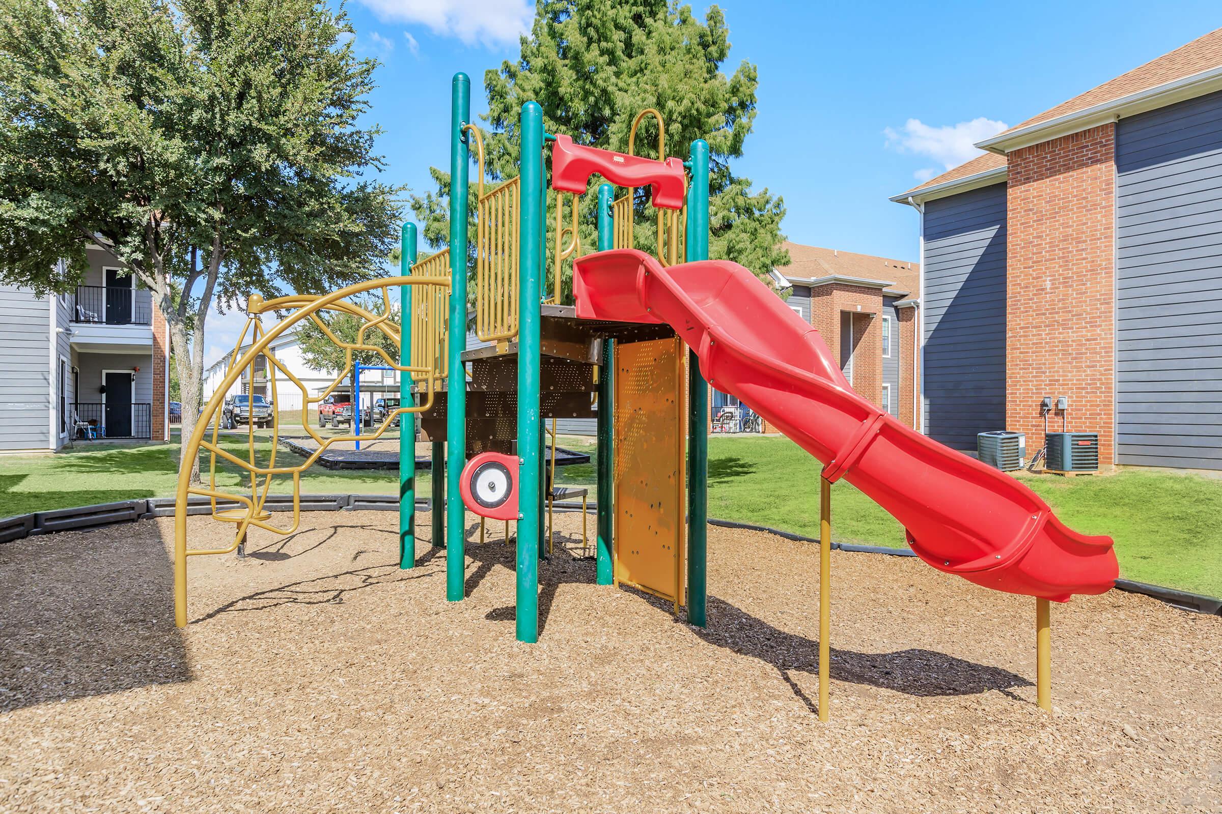 A colorful playground structure featuring a bright red slide, climbing bars, and a target panel, situated on a bed of mulch. Surrounding the playground are green grass and trees, with residential buildings visible in the background under a clear blue sky.