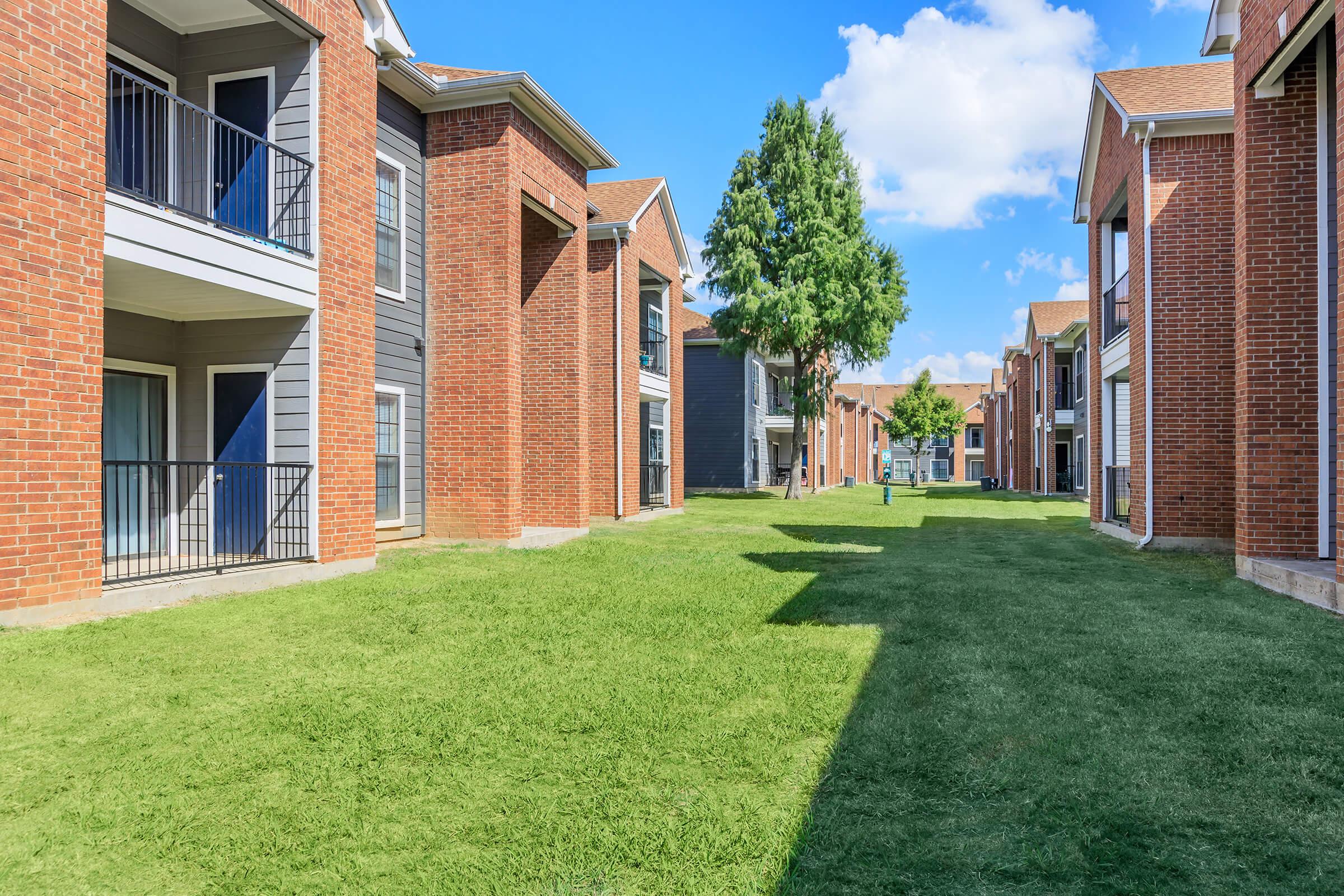 View of a landscaped apartment complex featuring two-story brick buildings, green lawns, and clear blue skies. The scene includes a pathway bordered by grass, with trees providing shade, creating a peaceful and inviting community atmosphere.