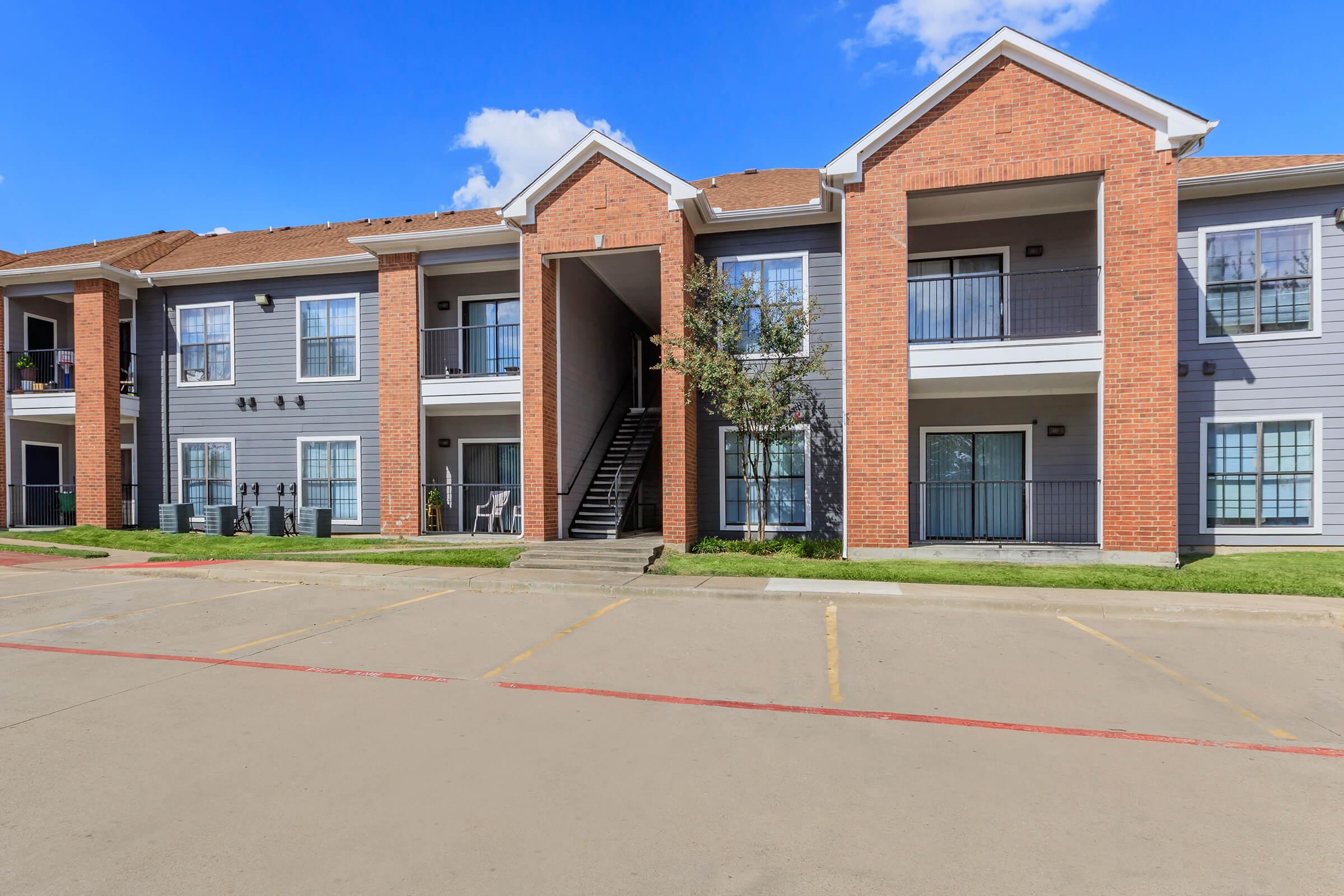 Modern apartment building with red brick façade and balcony. Two stories with stairs leading to the upper level. Green lawn in front and clear blue sky overhead. Parking area visible in the foreground.