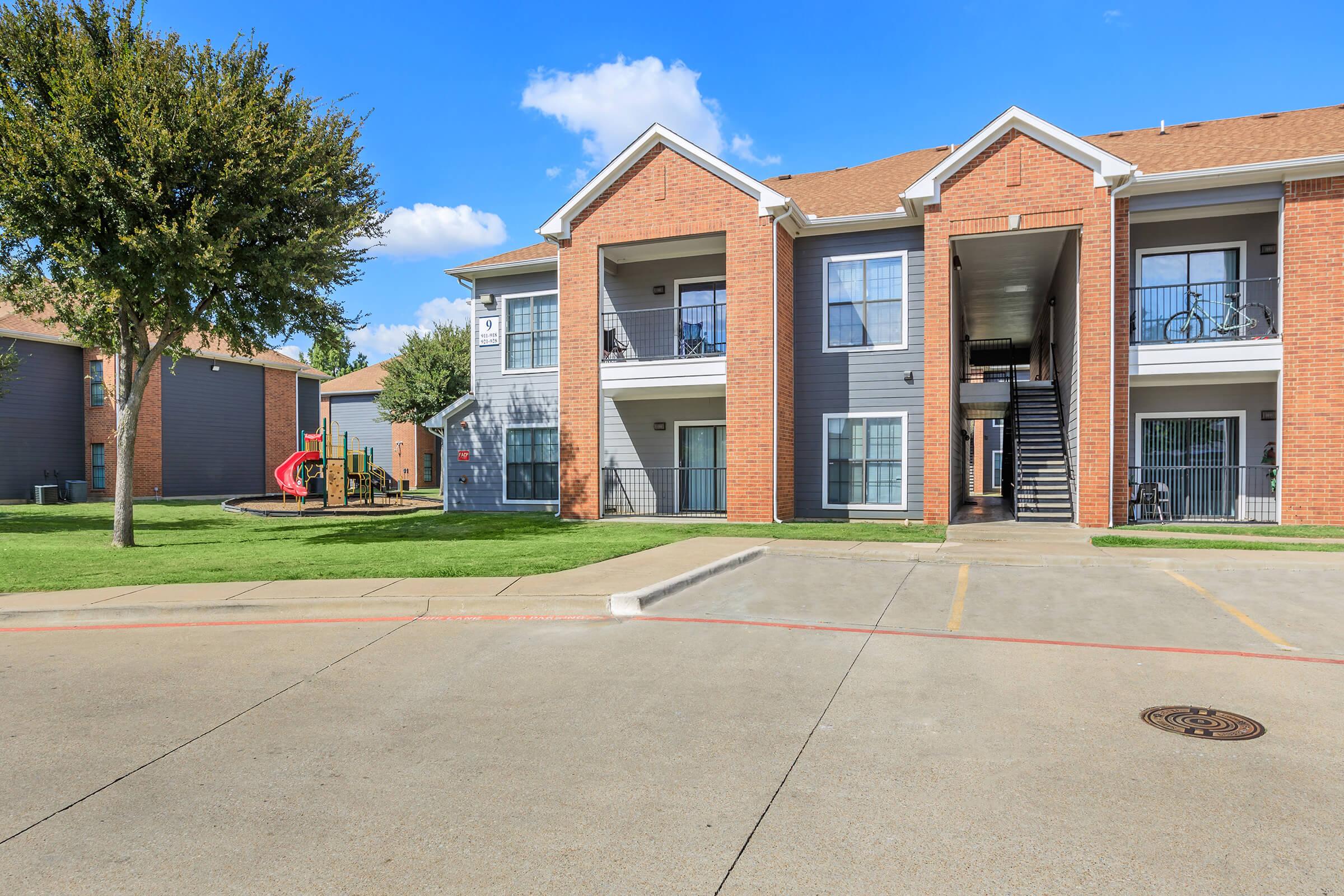 A multi-story apartment building with brick and gray siding, featuring balconies and a staircase. In the background, there's a playground with green grass and trees. The sky is clear with a few clouds, and the parking area is visible in the foreground.