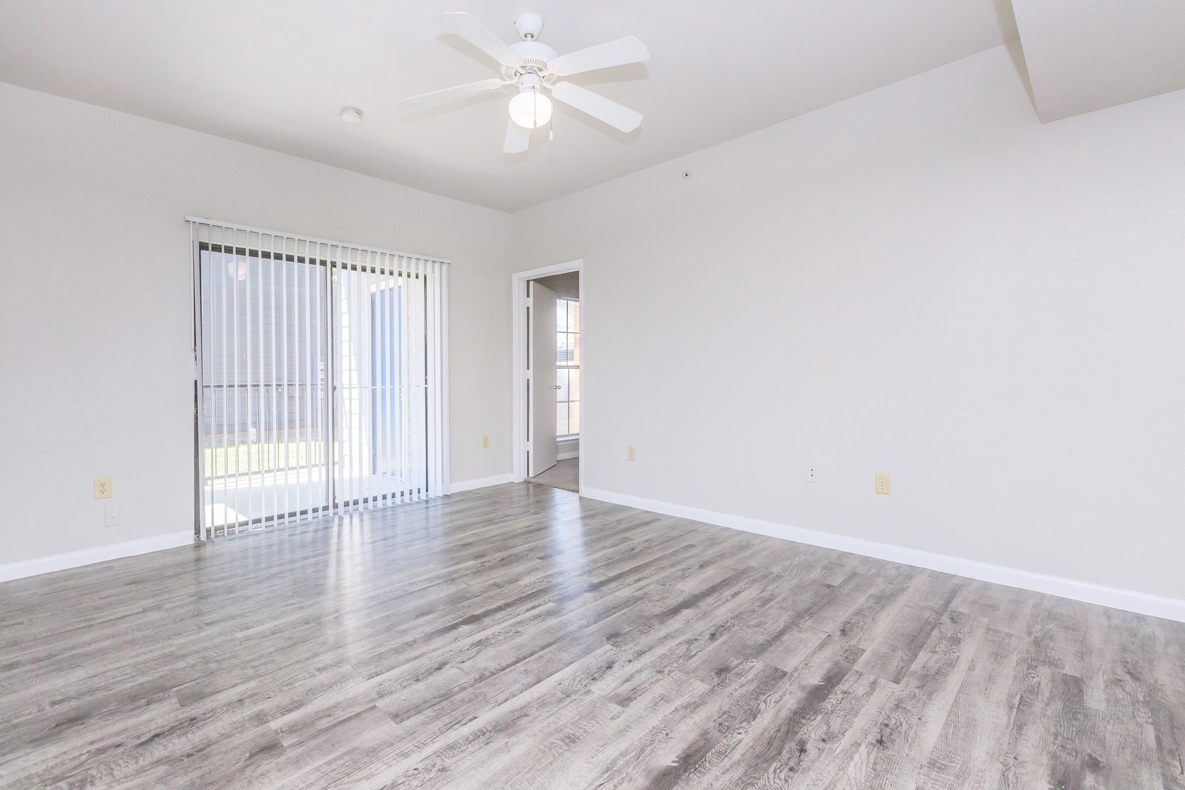 A spacious, empty room with light-colored walls and wood-look flooring. A ceiling fan is visible, and sliding glass doors provide natural light, leading to an outdoor area. There is a doorway on the right side, suggesting access to another room or hallway.