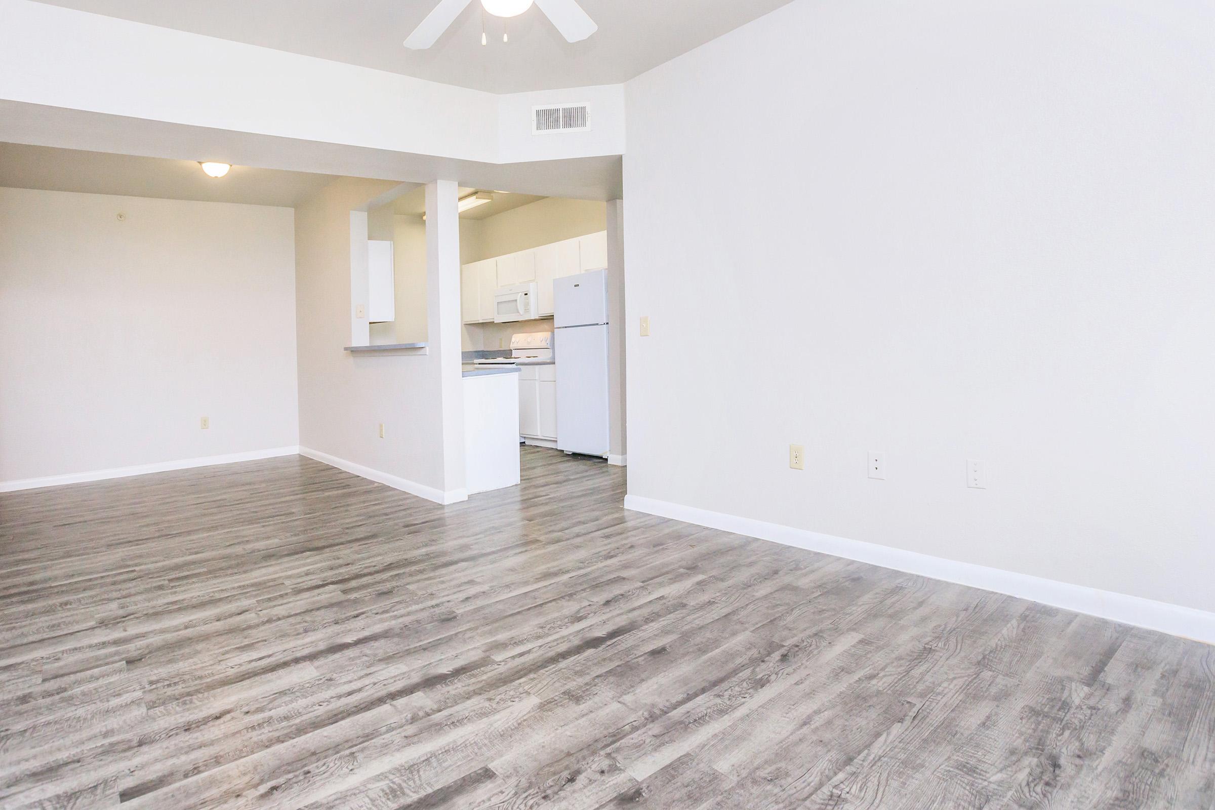 Spacious empty living area with light gray vinyl flooring, a ceiling fan, and white walls. A small kitchen area with white cabinets and appliances is visible in the background, including a refrigerator and stove. Natural light fills the room from a nearby window.