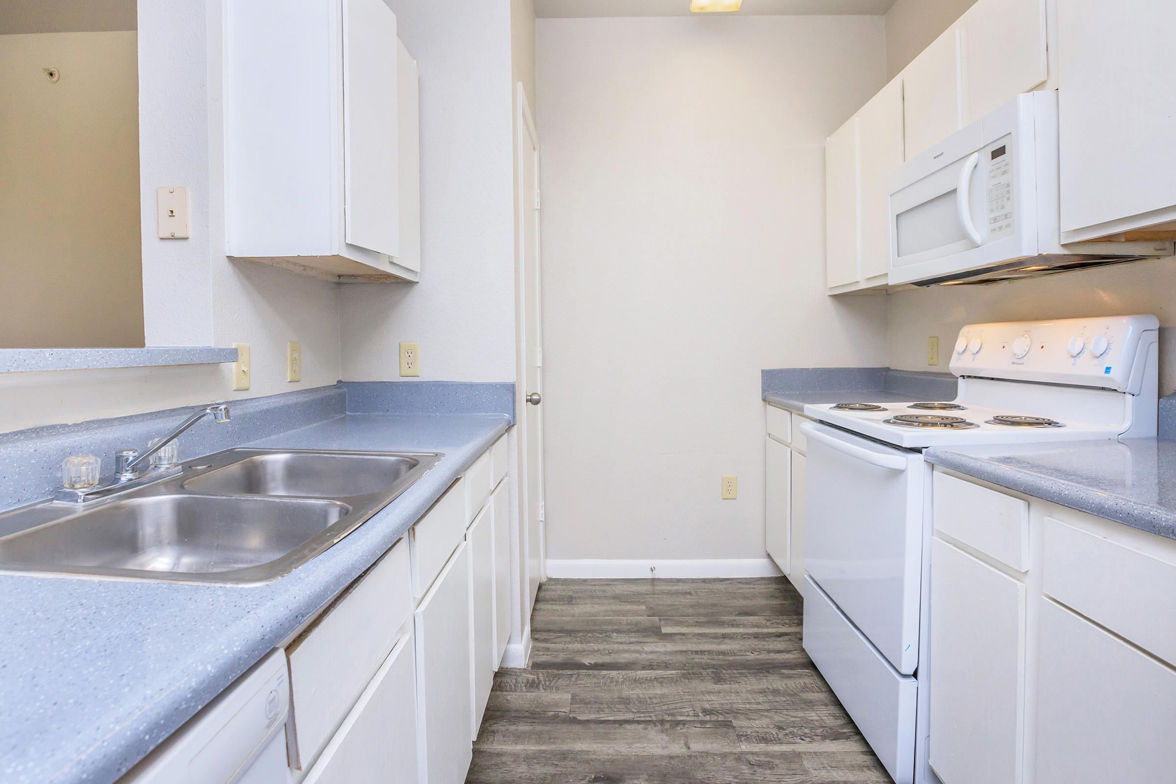 A clean, modern kitchen with white cabinets and countertops. The space features a double sink, a stove, an oven, and a microwave. Grey vinyl flooring complements the light-colored walls, creating a bright and airy atmosphere. The layout is functional, with ample storage and work space.
