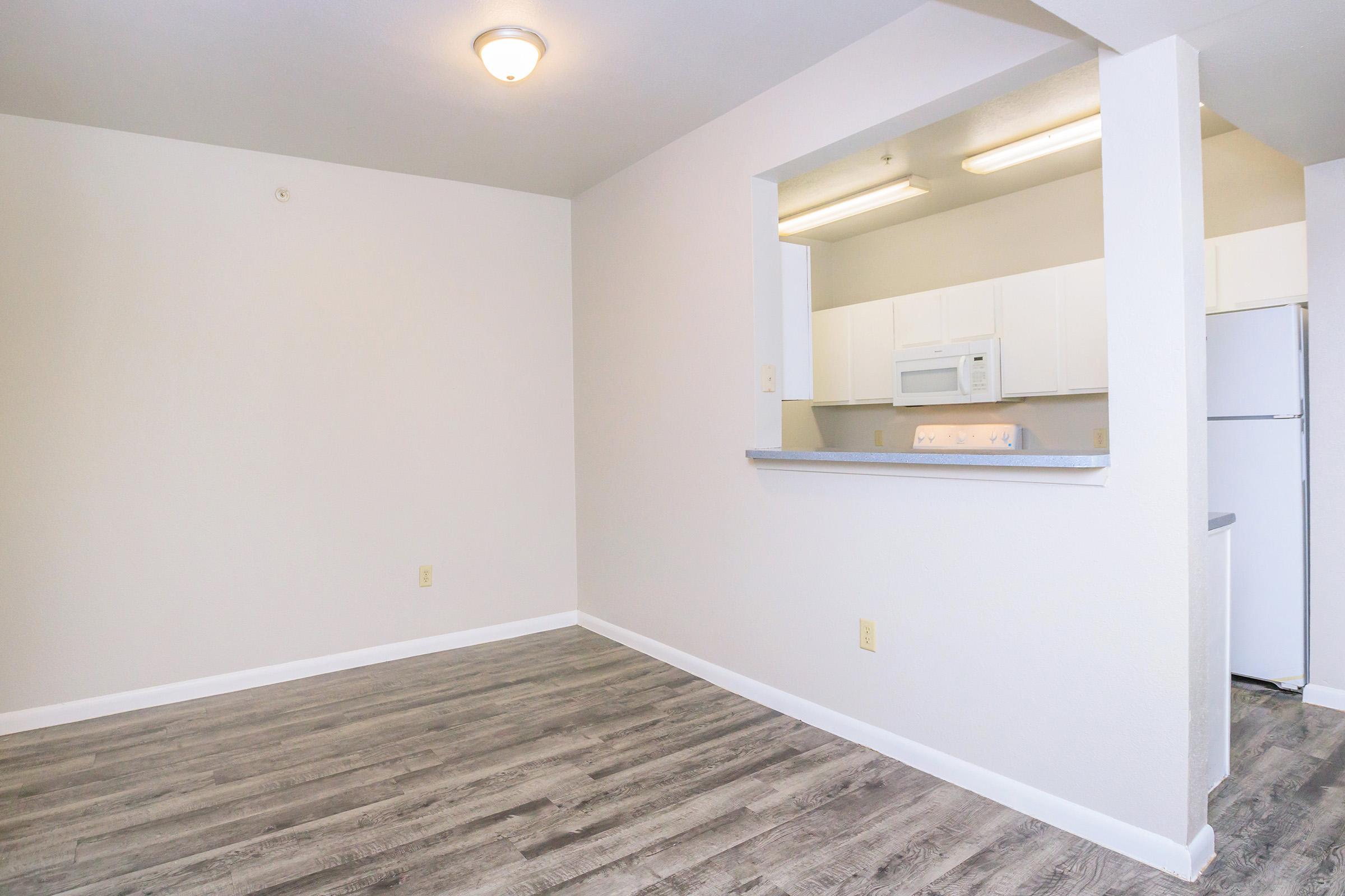 An empty apartment living area with light gray walls and a wood-like floor. There's a small kitchen visible through an opening in the wall, featuring white cabinets, a microwave, and a refrigerator. The lighting is bright, and the space appears clean and ready for furnishing.
