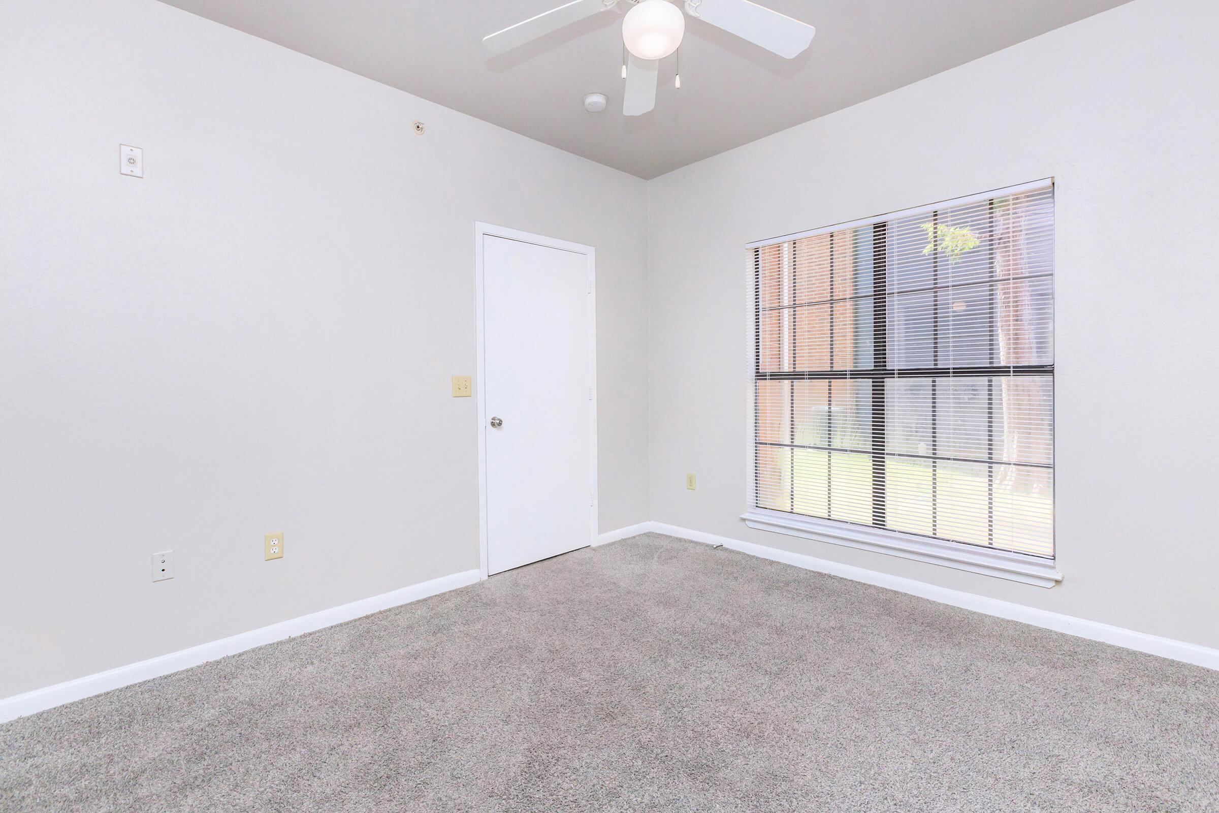 A spacious, empty room with light gray walls and beige carpet. It features a single ceiling fan, a white door, and a large window with blinds allowing natural light to enter. The room is unfurnished, creating a clean and open space.