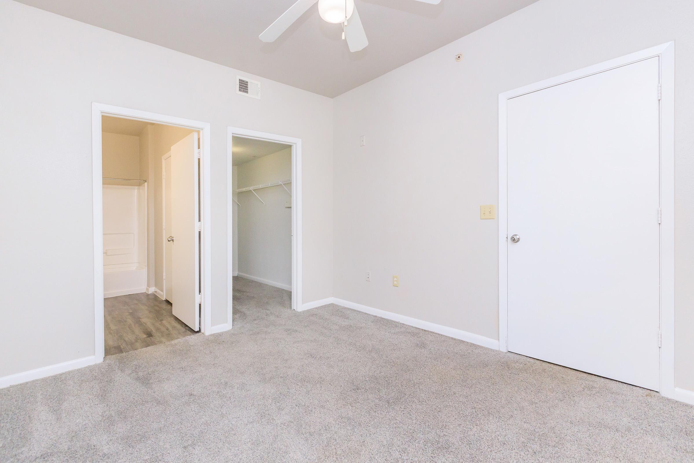 A well-lit room with neutral-colored walls and carpet flooring. The space features a ceiling fan and two doorways leading to different areas, including a closet. One doorway is partially open, revealing a small bathroom with a bathtub. The overall ambiance is clean and simple, ideal for a bedroom or living area.