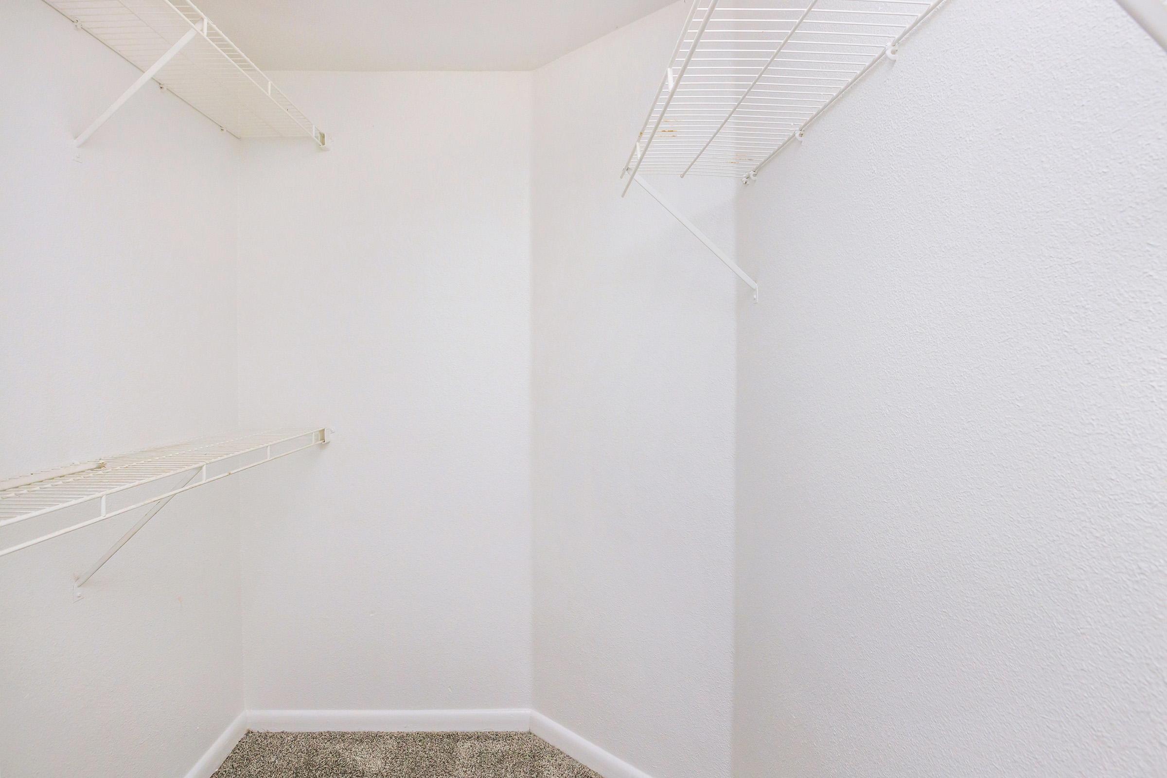 An empty closet with white walls and shelves. The shelves are made of wire and are mounted on two walls. The floor is covered with light-colored carpet, and there's a sense of spaciousness in the uncluttered space.