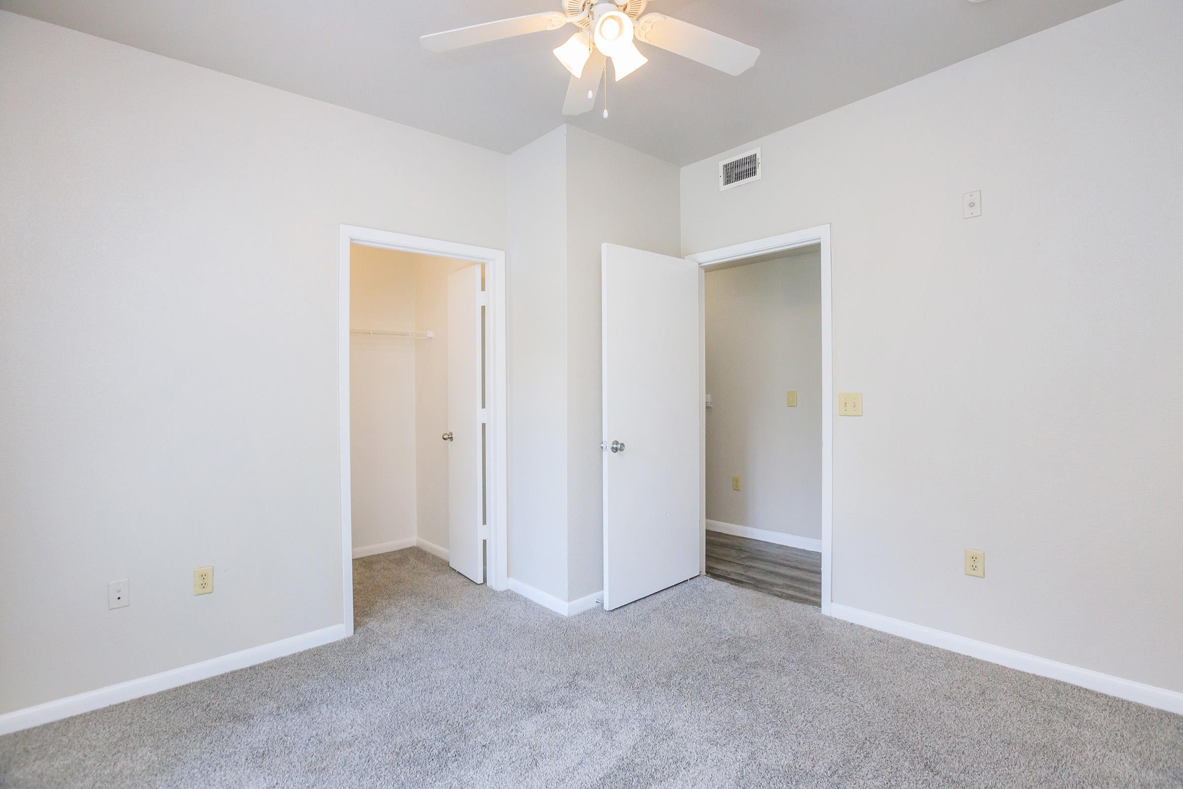 A well-lit empty room featuring light gray walls and carpet flooring. Two white doors are visible, one leading to a closet and the other to another room. A ceiling fan is installed above, and the overall space has a clean, modern aesthetic.