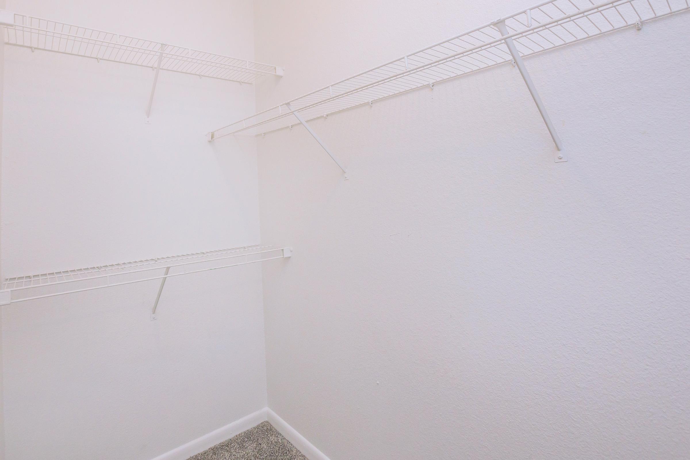 A clean, empty closet with white walls and two wire shelving units mounted on the walls. The shelves are arranged at different heights, with ample floor space below. The floor is carpeted in a light color, providing a simple and uncluttered appearance.