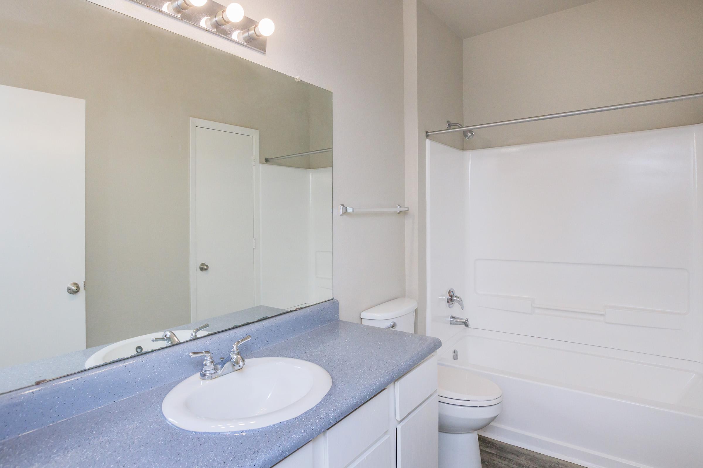 A modern bathroom featuring a sink with a countertop, a large mirror, a shower/tub combination with a white surround, and a toilet. The walls are painted light colors, and the space is well-lit with overhead lighting. The floor has a textured appearance, adding to the overall clean look.