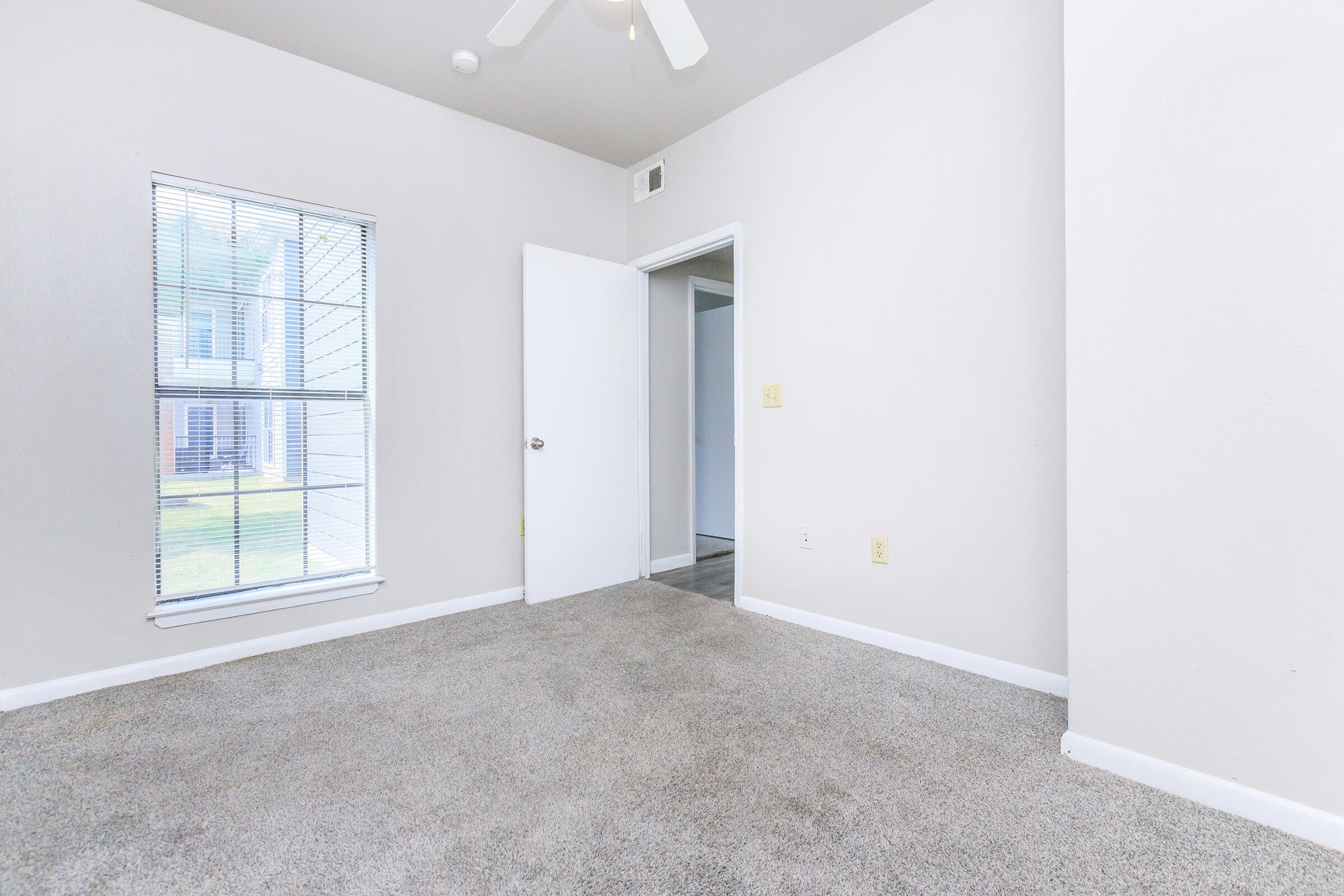 A bright, empty room with light gray walls and carpet, featuring a window with blinds letting in natural light. There is a ceiling fan and an open door leading to another room. The room appears spacious and clean, suitable for various purposes.