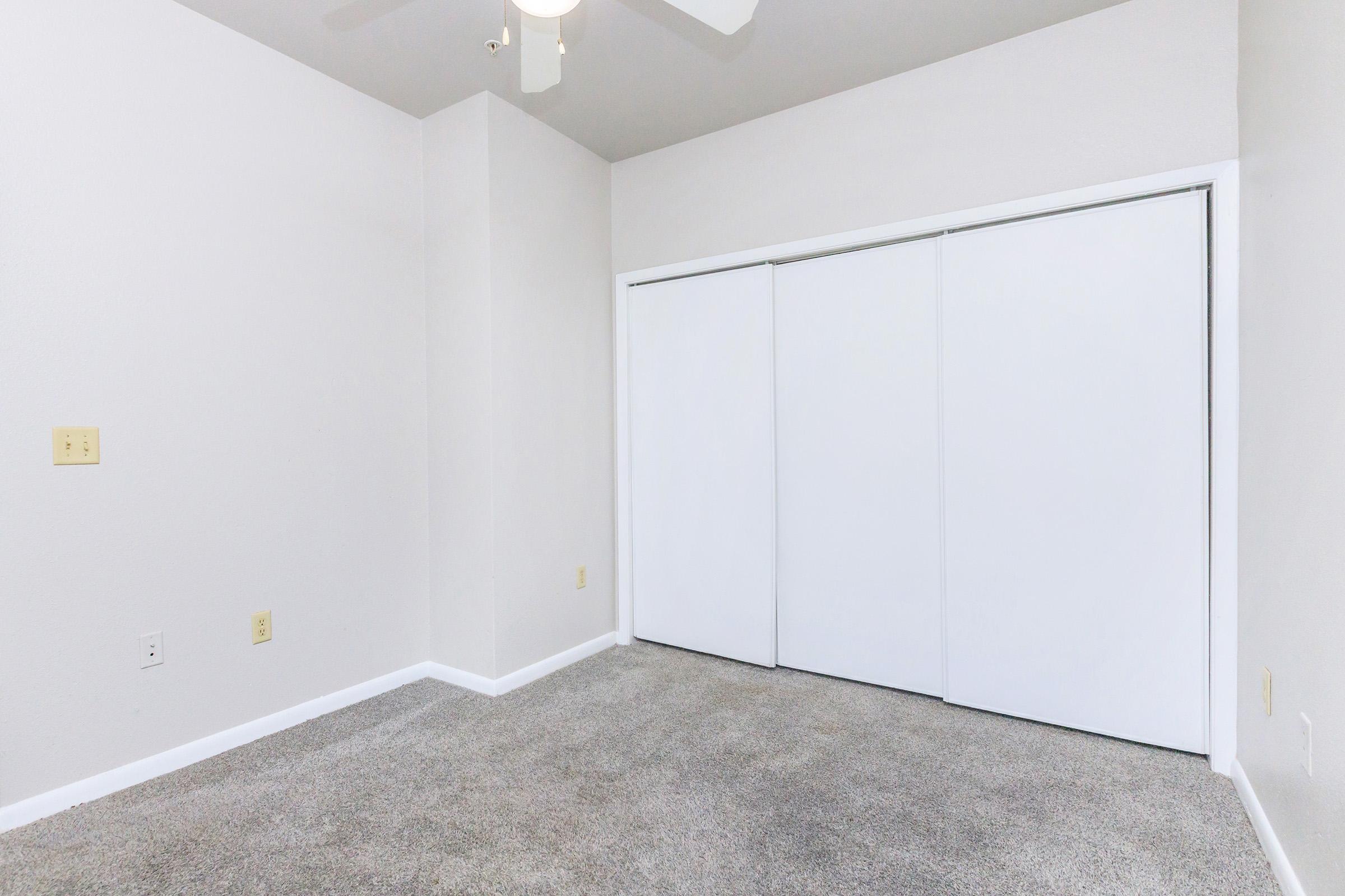 A clean, empty room with light gray carpeting and white walls. There is a ceiling fan and a large closet with sliding white doors. The space appears bright and unadorned, suitable for personalization or decoration.