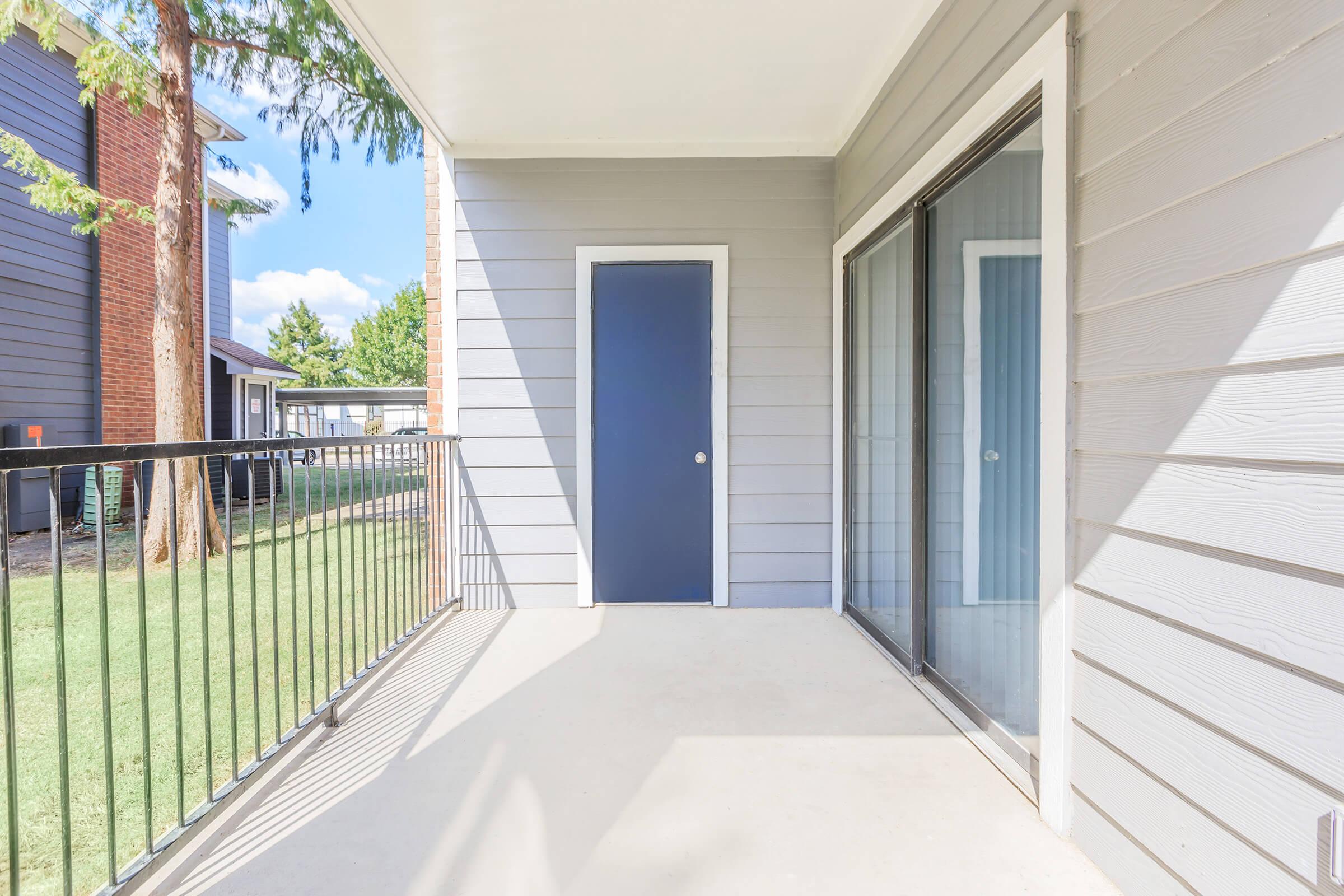 A view of a porch area with a beige concrete floor, a blue door, and sliding glass doors that lead to an indoor space. The walls are gray, and there is a black railing, with green grass and trees visible in the background. Bright daylight illuminates the scene.