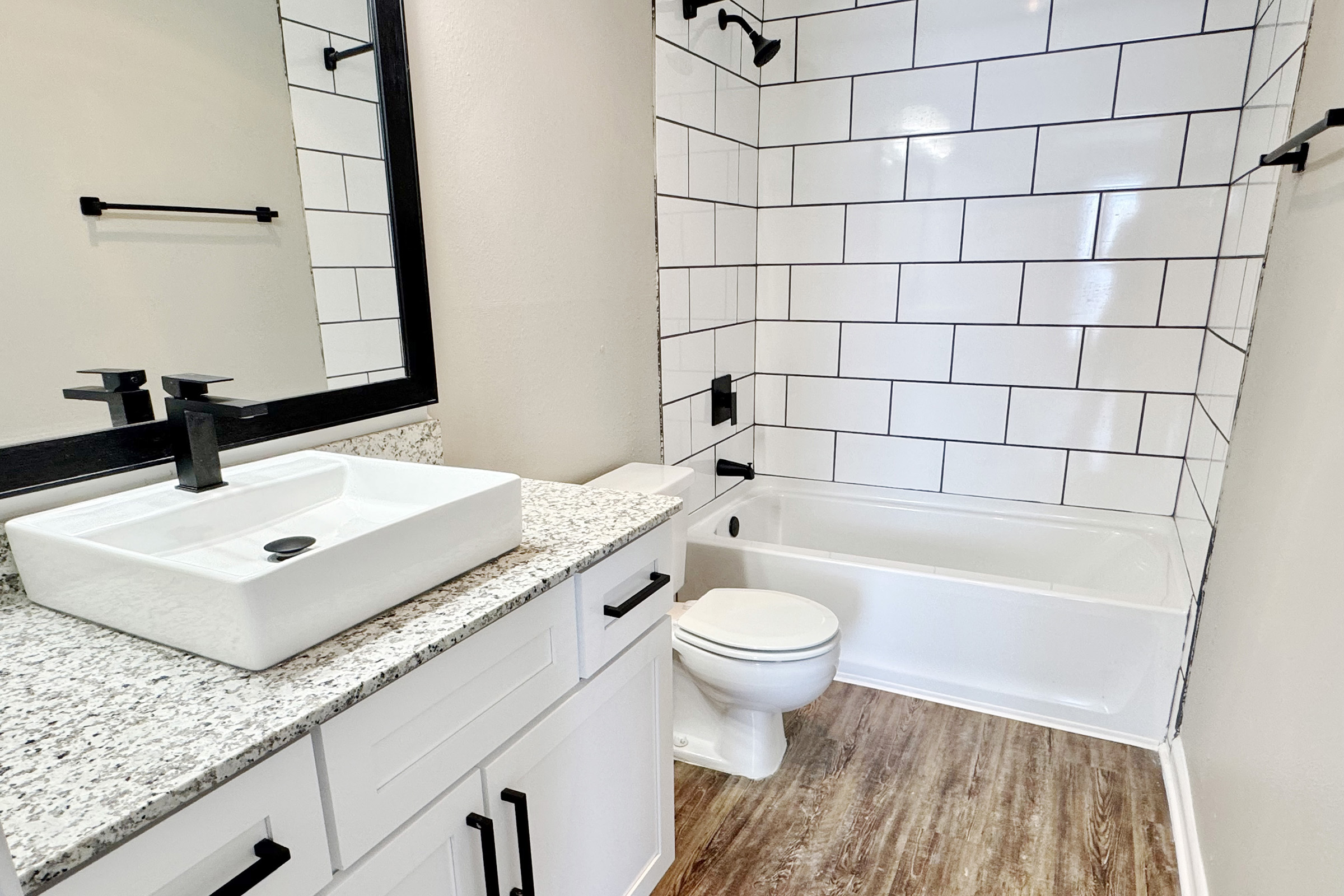 Modern bathroom featuring a white bathtub and shower combo with black fixtures, a large rectangular mirror, and a granite countertop with a vessel sink. The walls are tiled with white and black accents, and the flooring is a wood-like laminate.