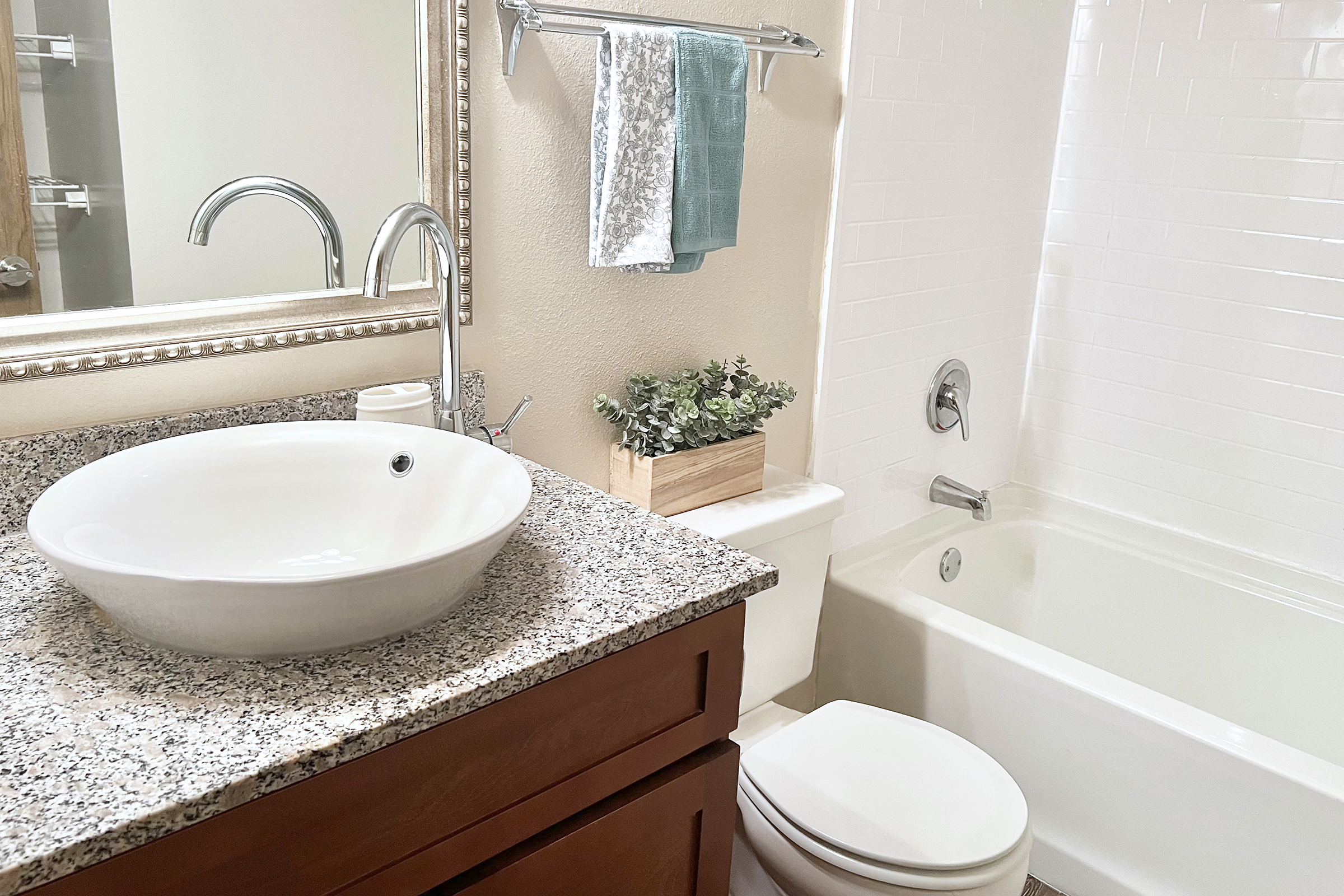 A well-lit bathroom featuring a modern white vessel sink on a granite countertop, a mirror above the sink, a towel rack with a few towels, a small plant in a wooden planter, and a bathtub next to a white toilet. Neutral-colored walls and tiles create a clean, inviting atmosphere.