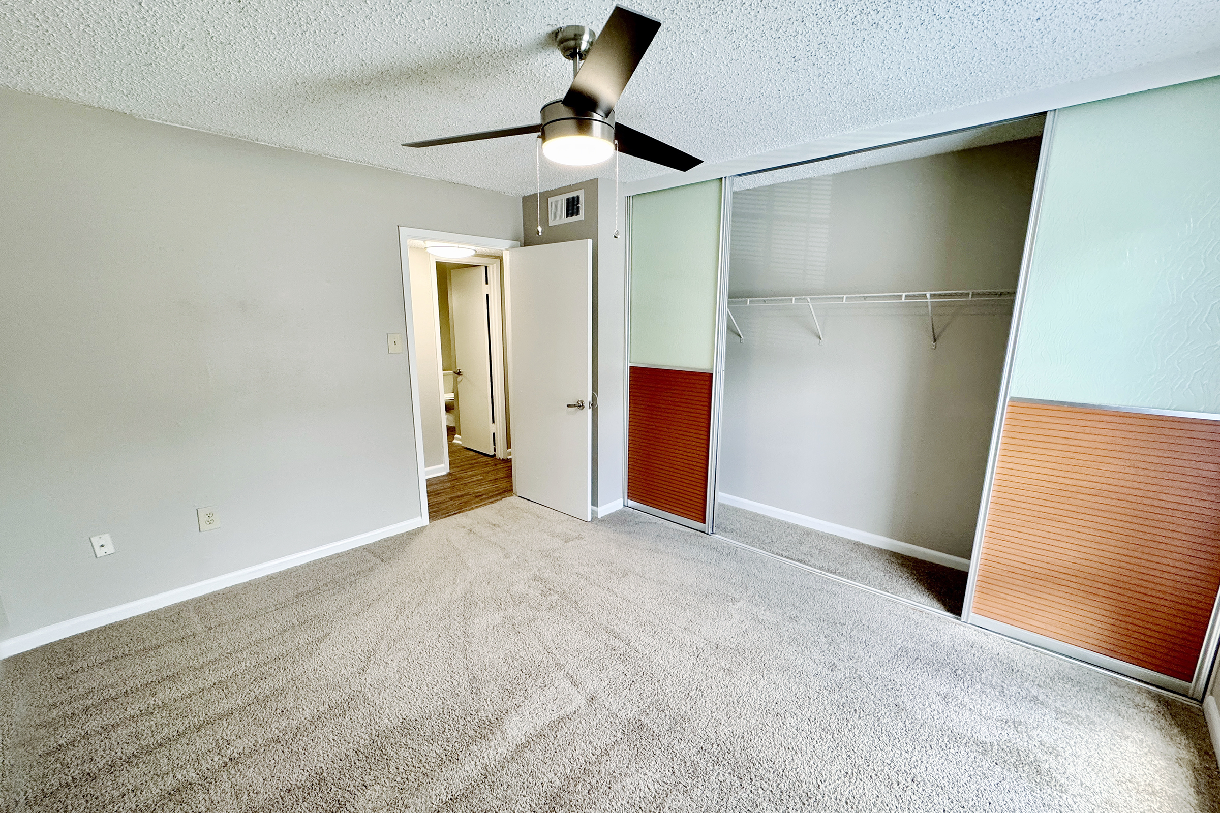 A spacious, well-lit bedroom featuring neutral-colored walls, a light ceiling fan, and plush carpet. On one side, there is a mirrored closet with sliding doors and an open area. A doorway in the background leads to a bathroom or hallway.