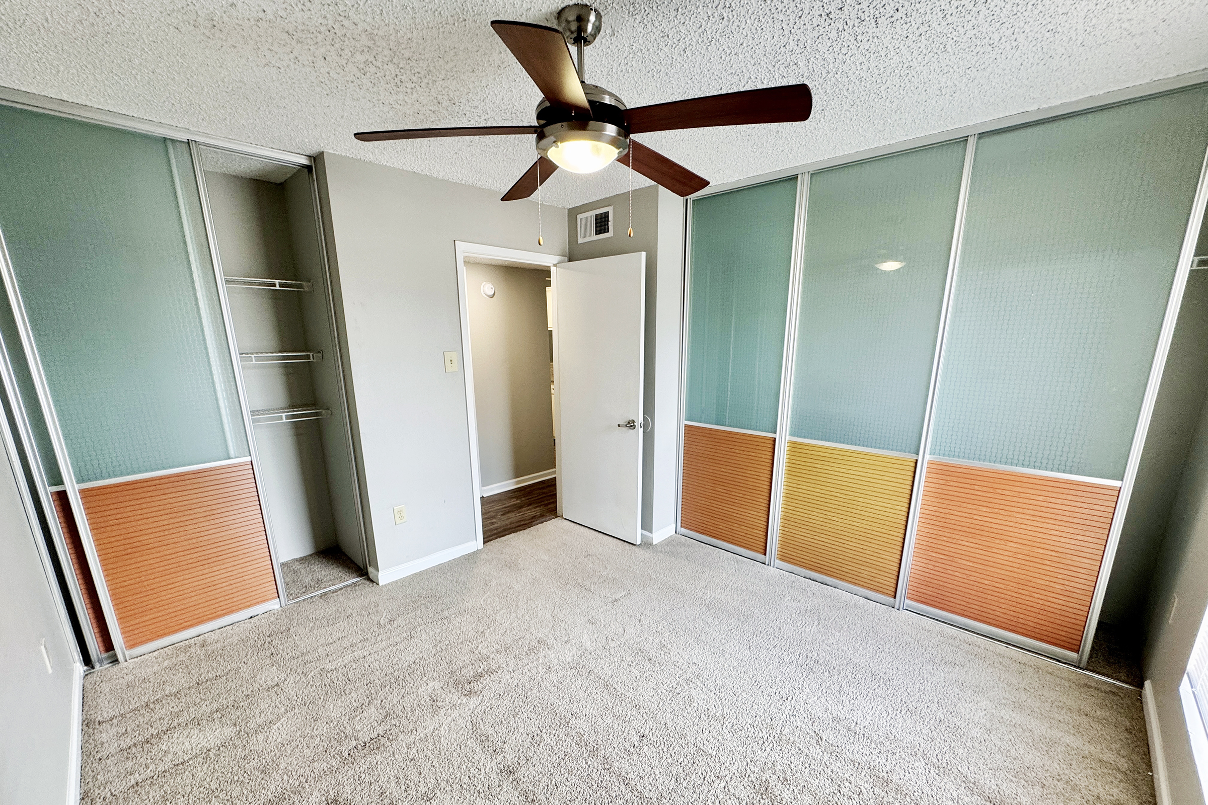 Empty bedroom featuring a ceiling fan, beige carpet, and two sliding closet doors with a combination of frosted glass and orange panels. A door leads to another area, and the walls are painted light gray. Natural light is coming from an unseen window.