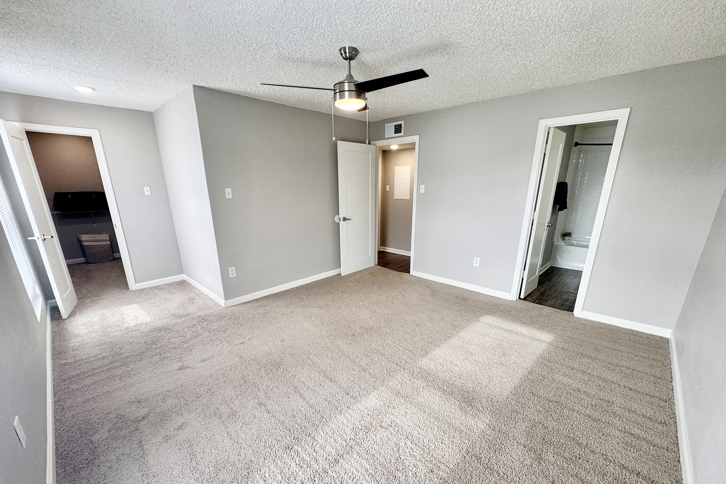 A spacious, well-lit room with beige carpet and light gray walls. A ceiling fan is installed, and there are two doors leading to adjacent areas. The room appears freshly painted and organized, creating a clean and inviting atmosphere. Natural light brightens the space from windows or doors.