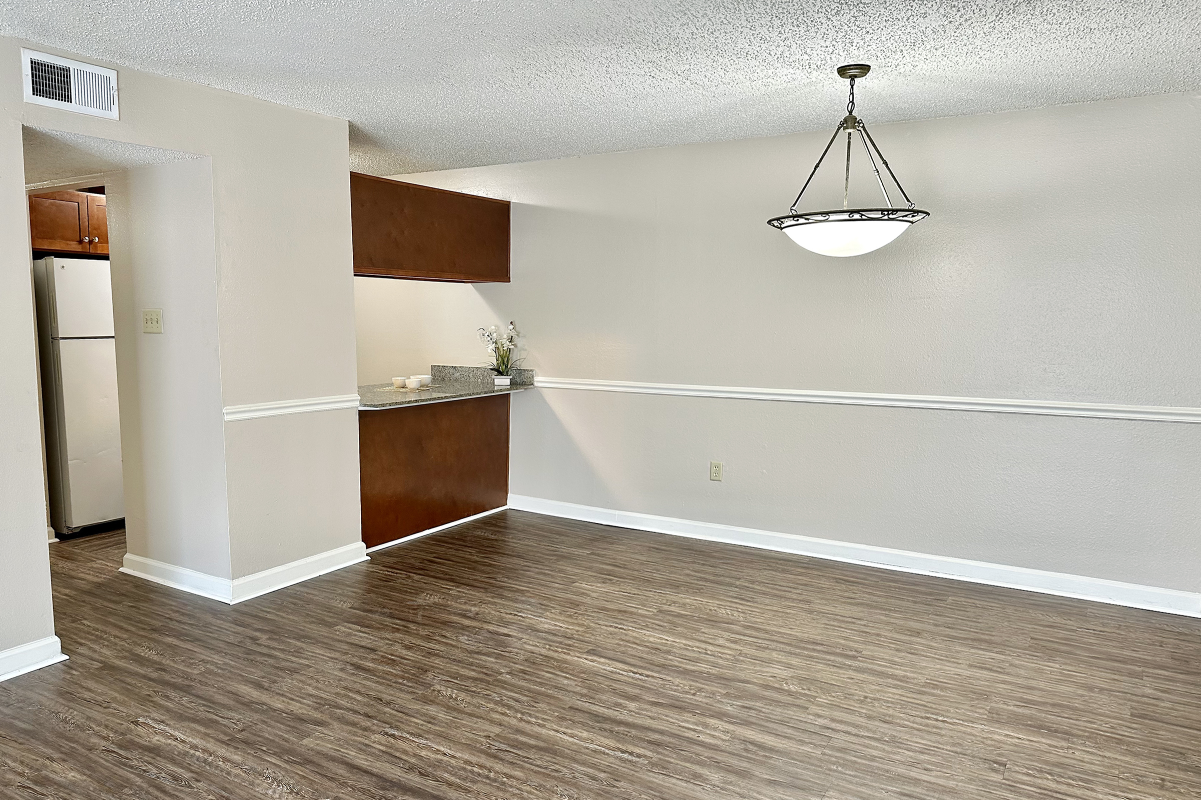 A spacious, well-lit living area with laminate flooring and light beige walls. To the left, a kitchen is partially visible, featuring dark wood cabinets and a white refrigerator. A decorative light fixture hangs from the ceiling. The room is empty, creating a minimalist and inviting atmosphere.