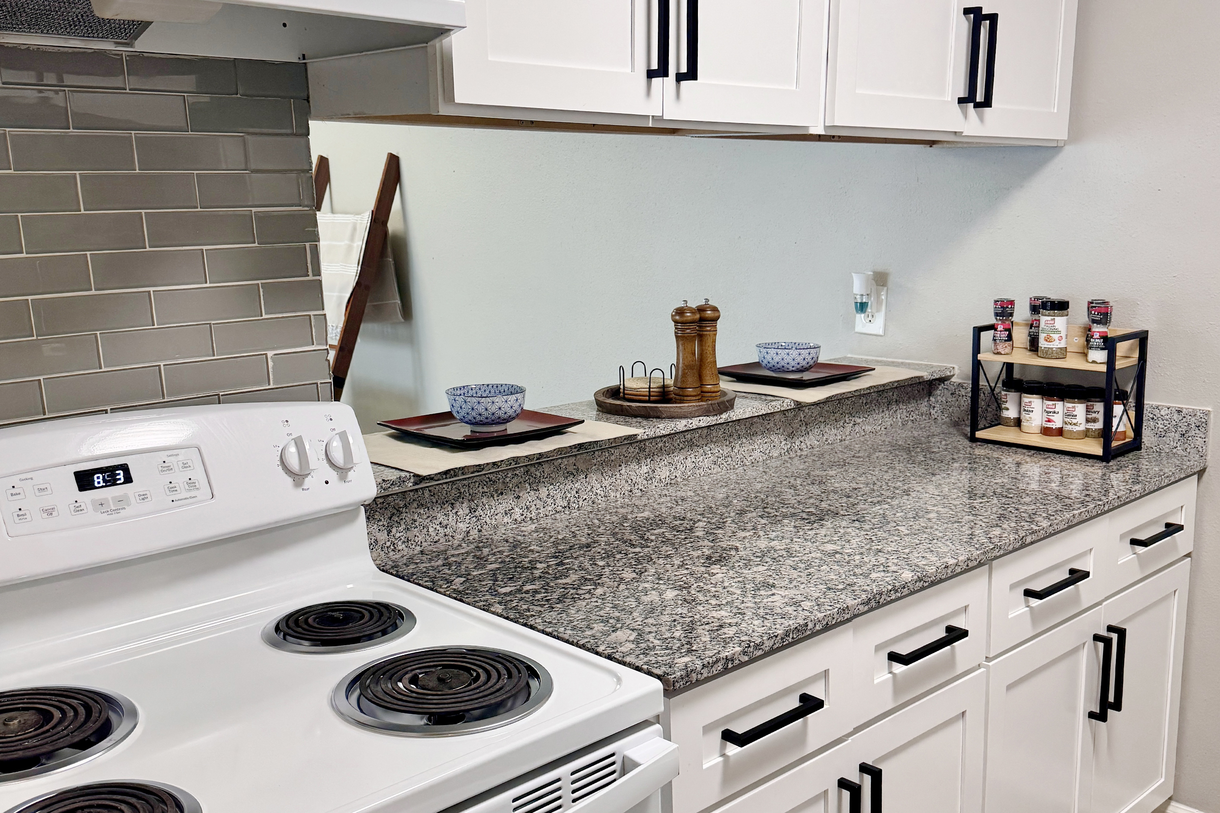 Modern kitchen countertop featuring a white stove, gray backsplash, and granite surface. Stylish decor includes a wooden pepper grinder, two blue bowls, and a spice rack with various spices, all set against light-colored walls and minimalistic cabinetry.